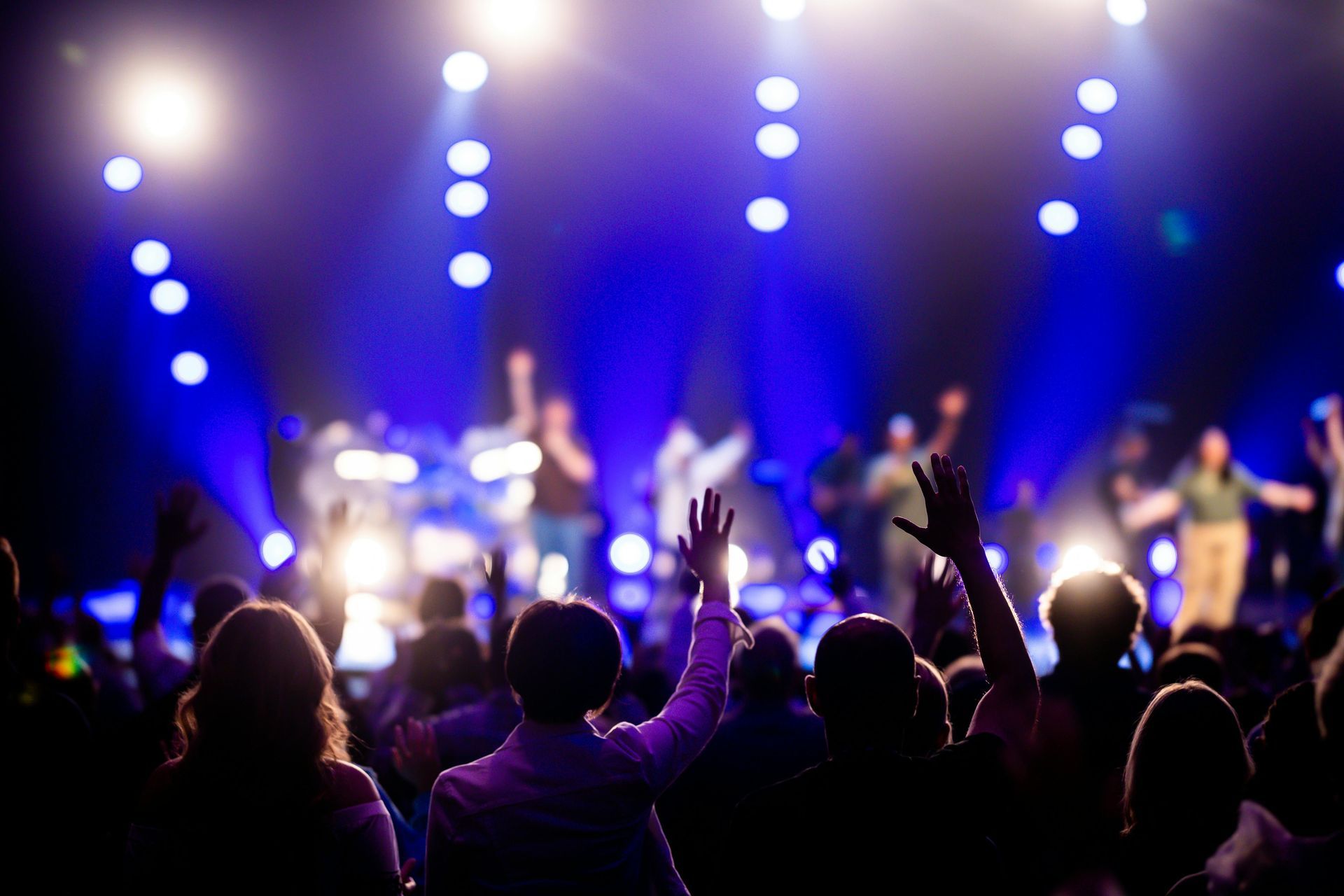 A concert audience stands with hands raised in the air, silhouetted against bright blue stage lighting.