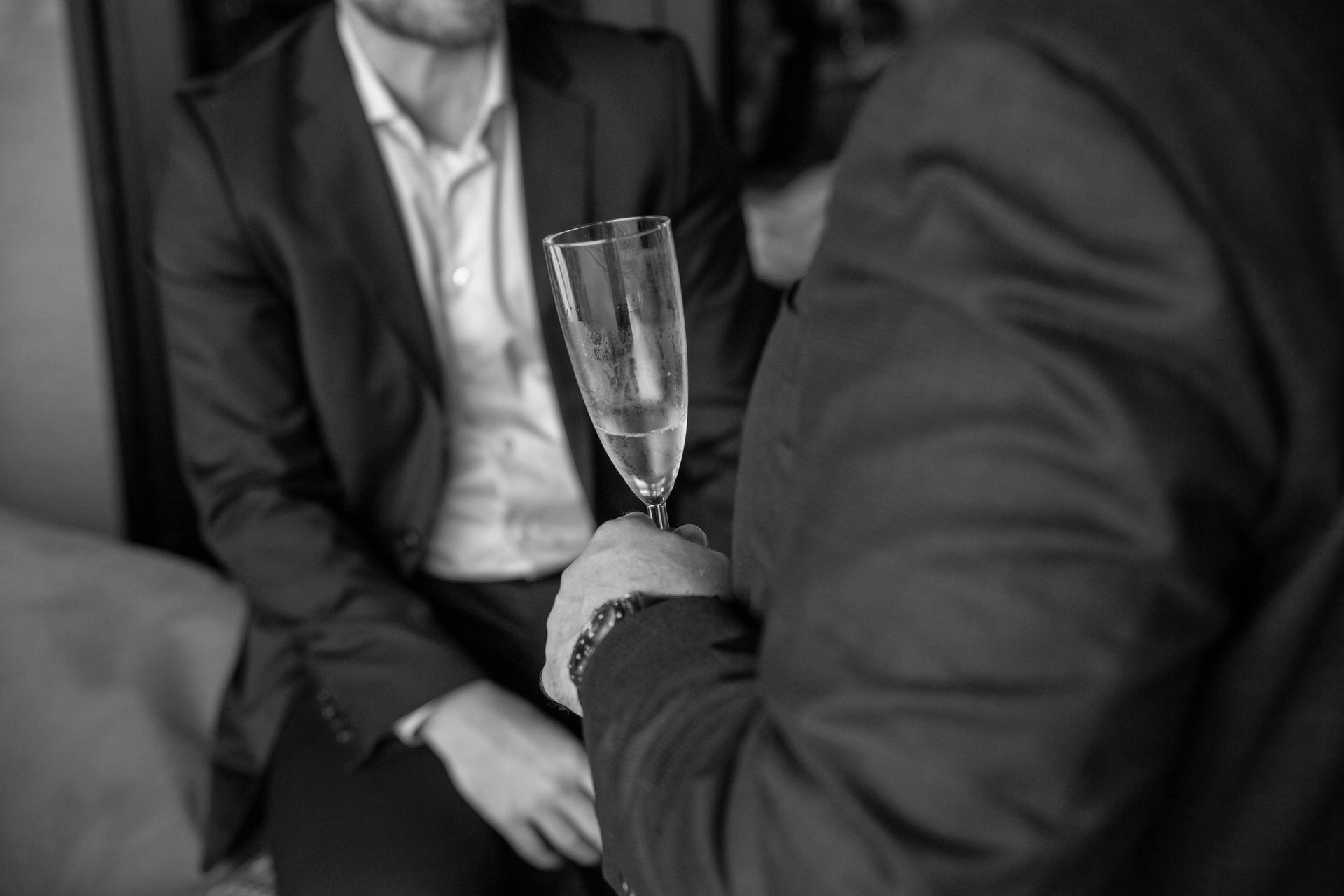 Two men in suits, one holding up a champagne flute, other seated. Black and white.