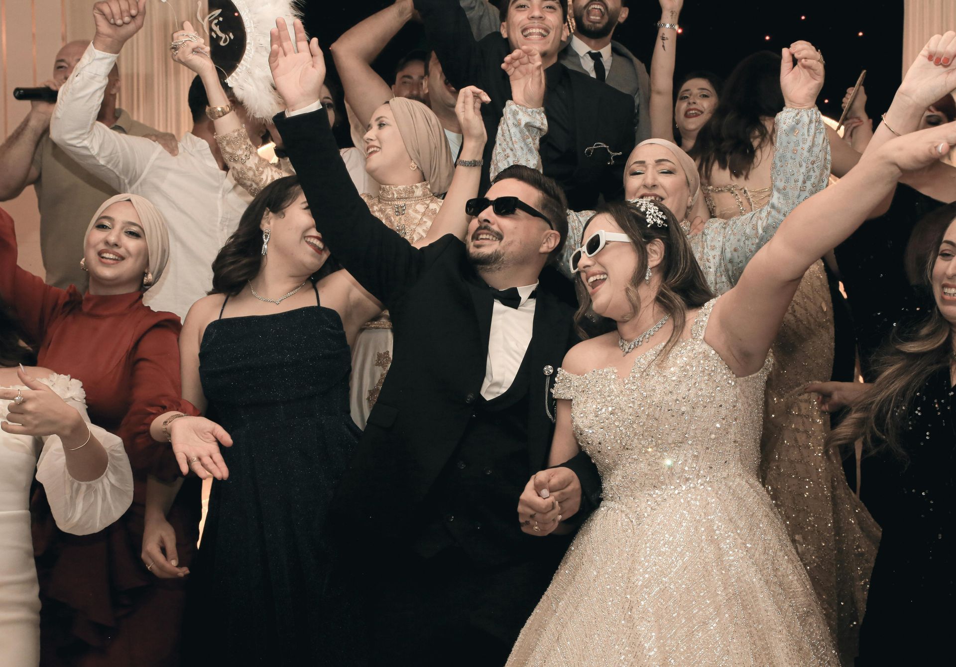A group of people celebrating at an event, with the central couple wearing sunglasses and dancing with arms raised.