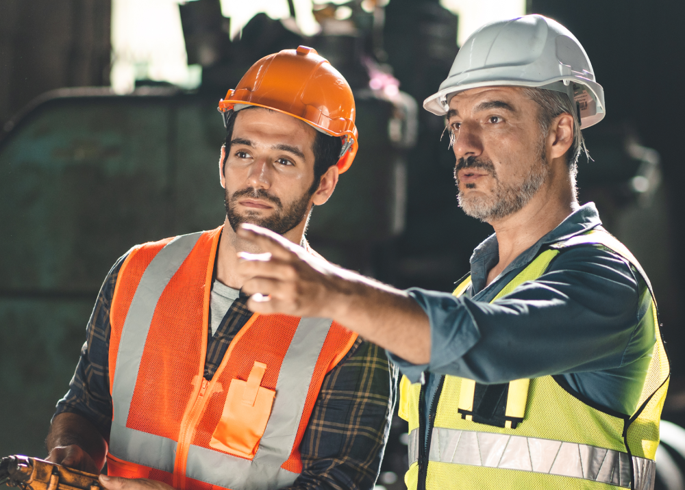 Two construction workers are standing next to each other and one of them is pointing at something.