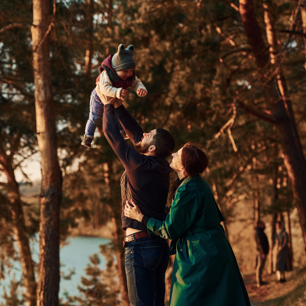 A man and woman are holding a baby in the air in the woods.
