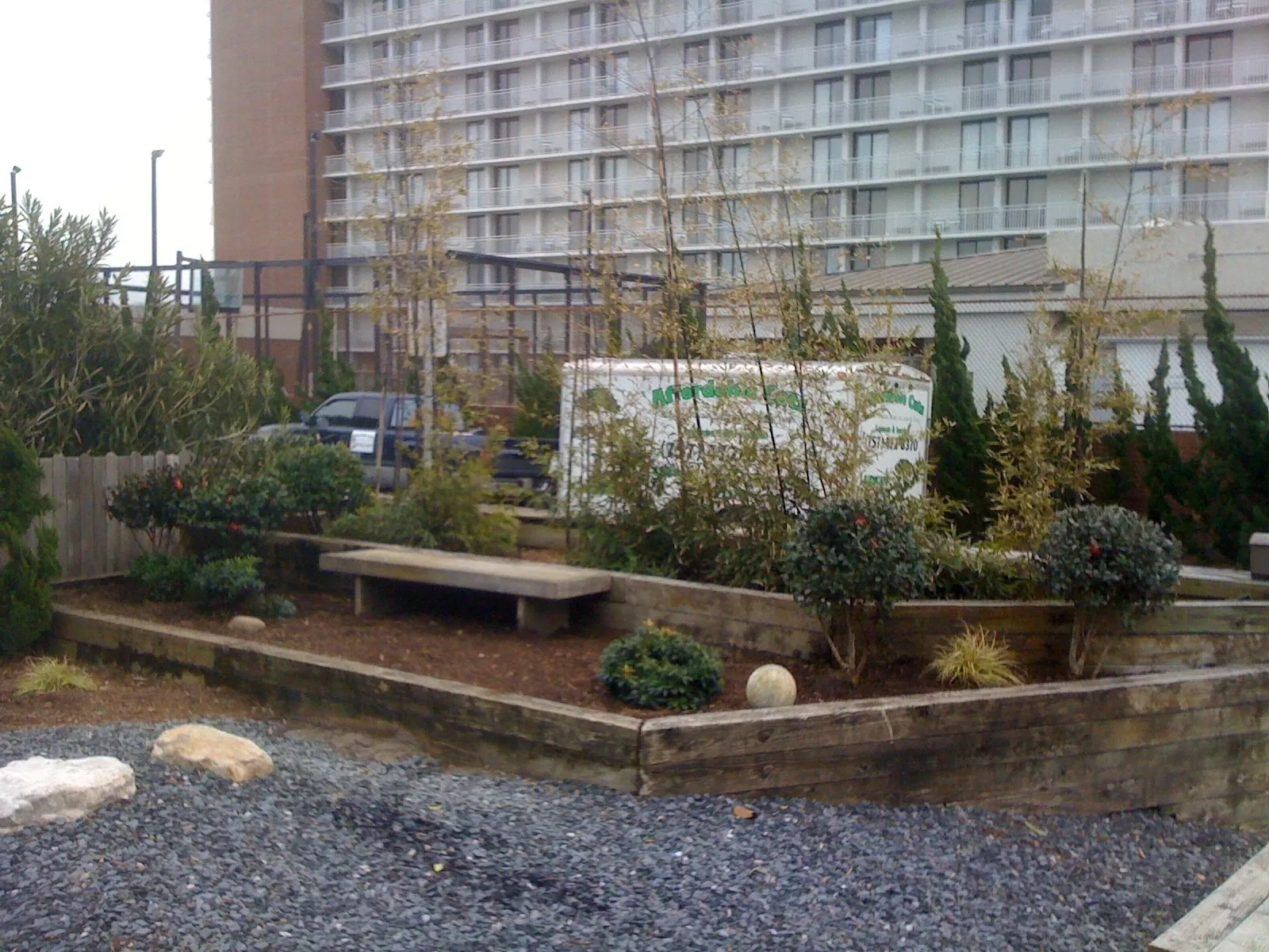 Raised garden bed with bench, small trees, and bushes in front of a multi-story building.
