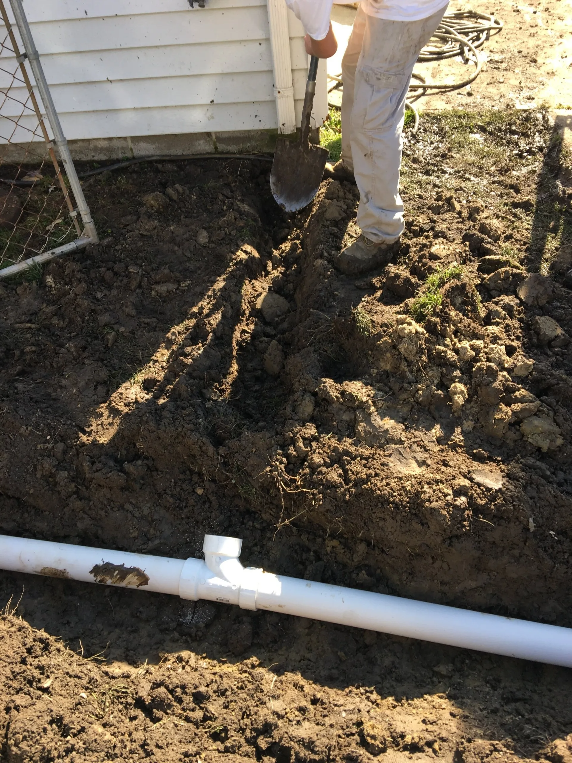 Person digging in dirt near a white PVC pipe and a building.