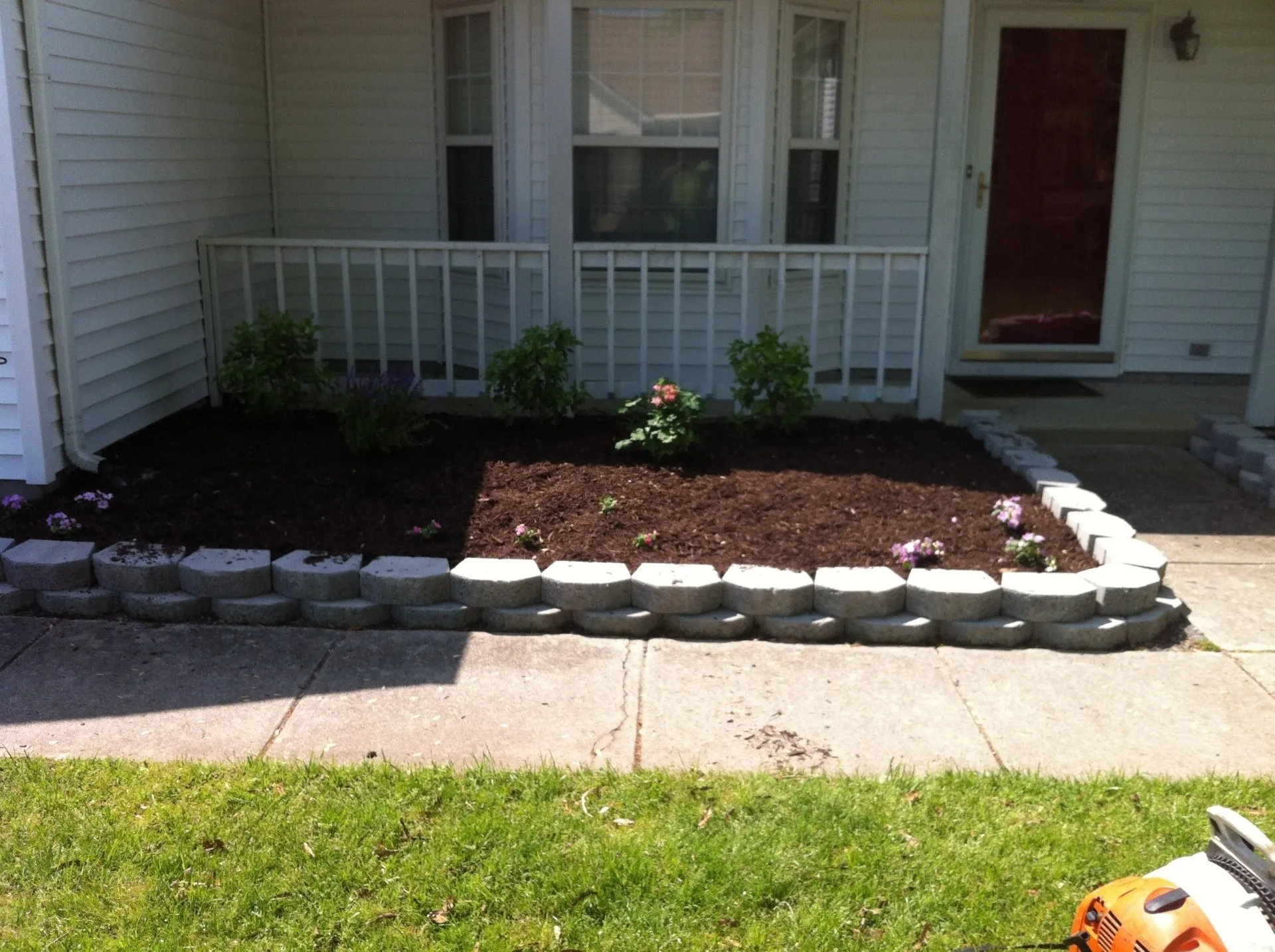 A house front yard with a flower bed, grey blocks border dark soil and small plants, and a lawn.