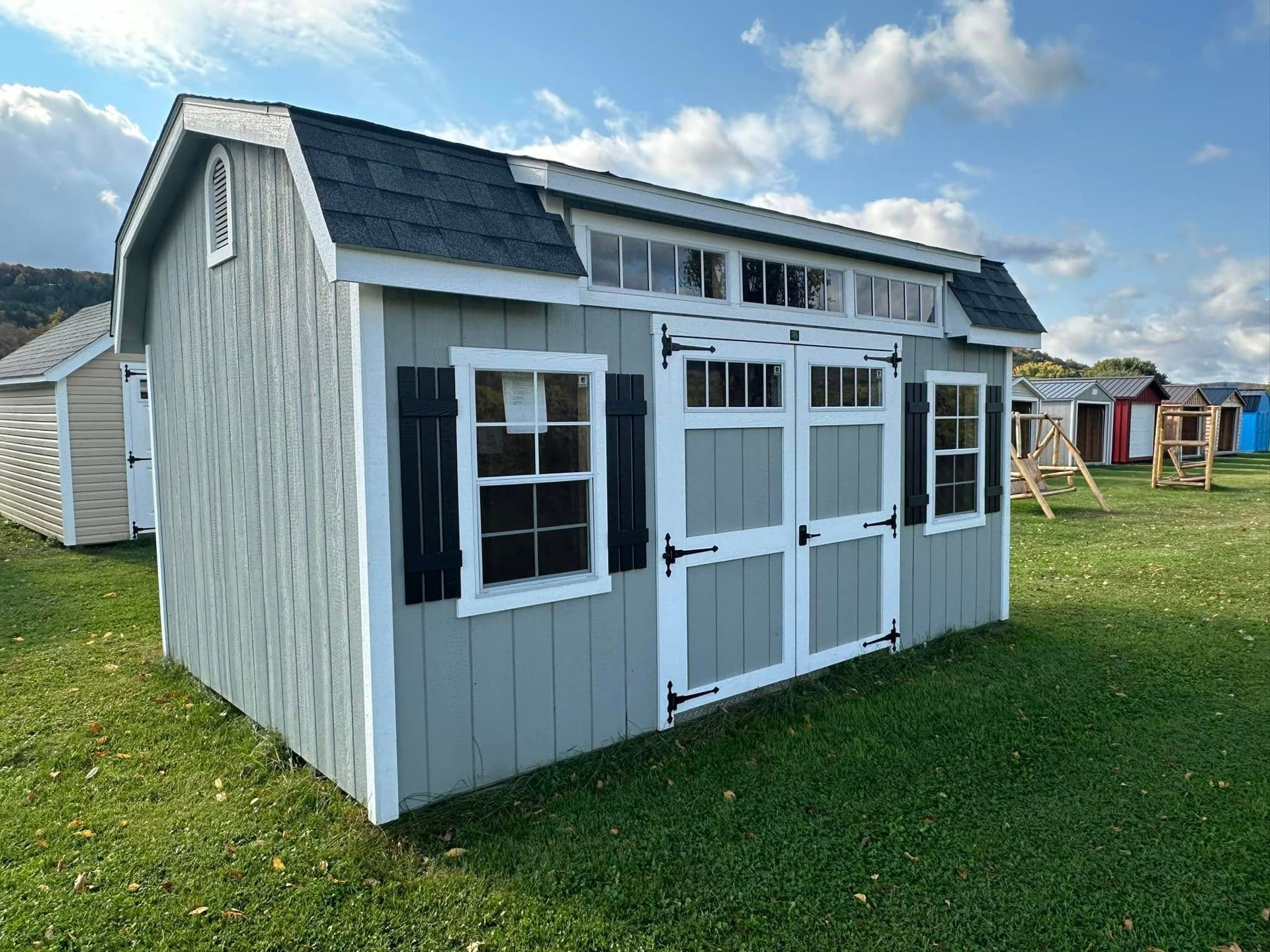 A gray and white shed with black shutters is sitting on top of a lush green field.