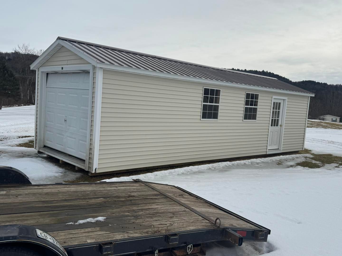 A small white garage is sitting on top of a snow covered field next to a tow truck.