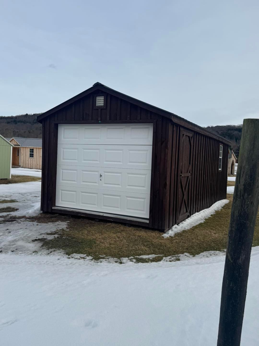 A wooden garage with a white garage door is sitting in the middle of a snowy field.