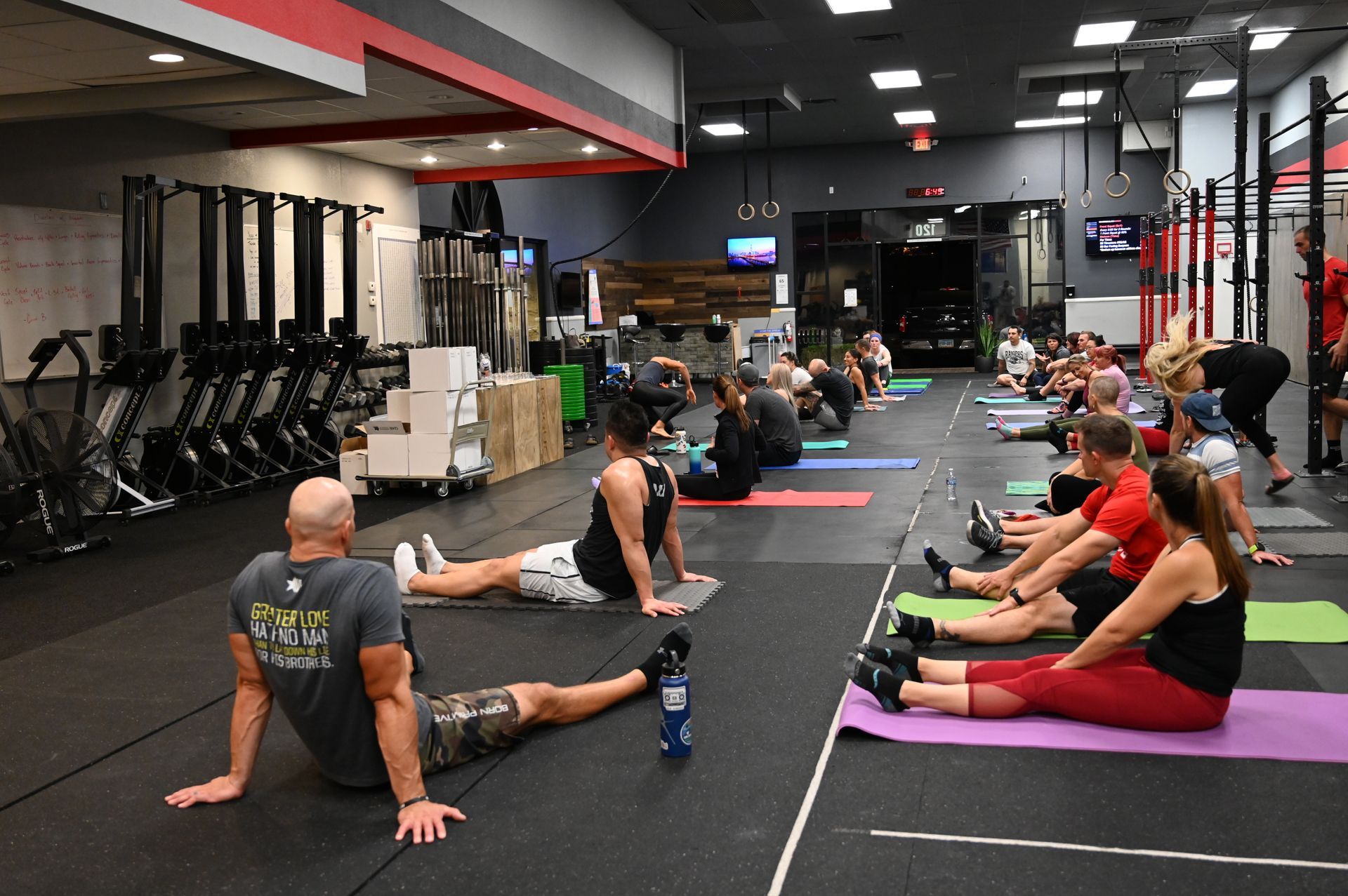 People stretching on mats at a gym, some with an instructor. Rows of exercise equipment line the walls.