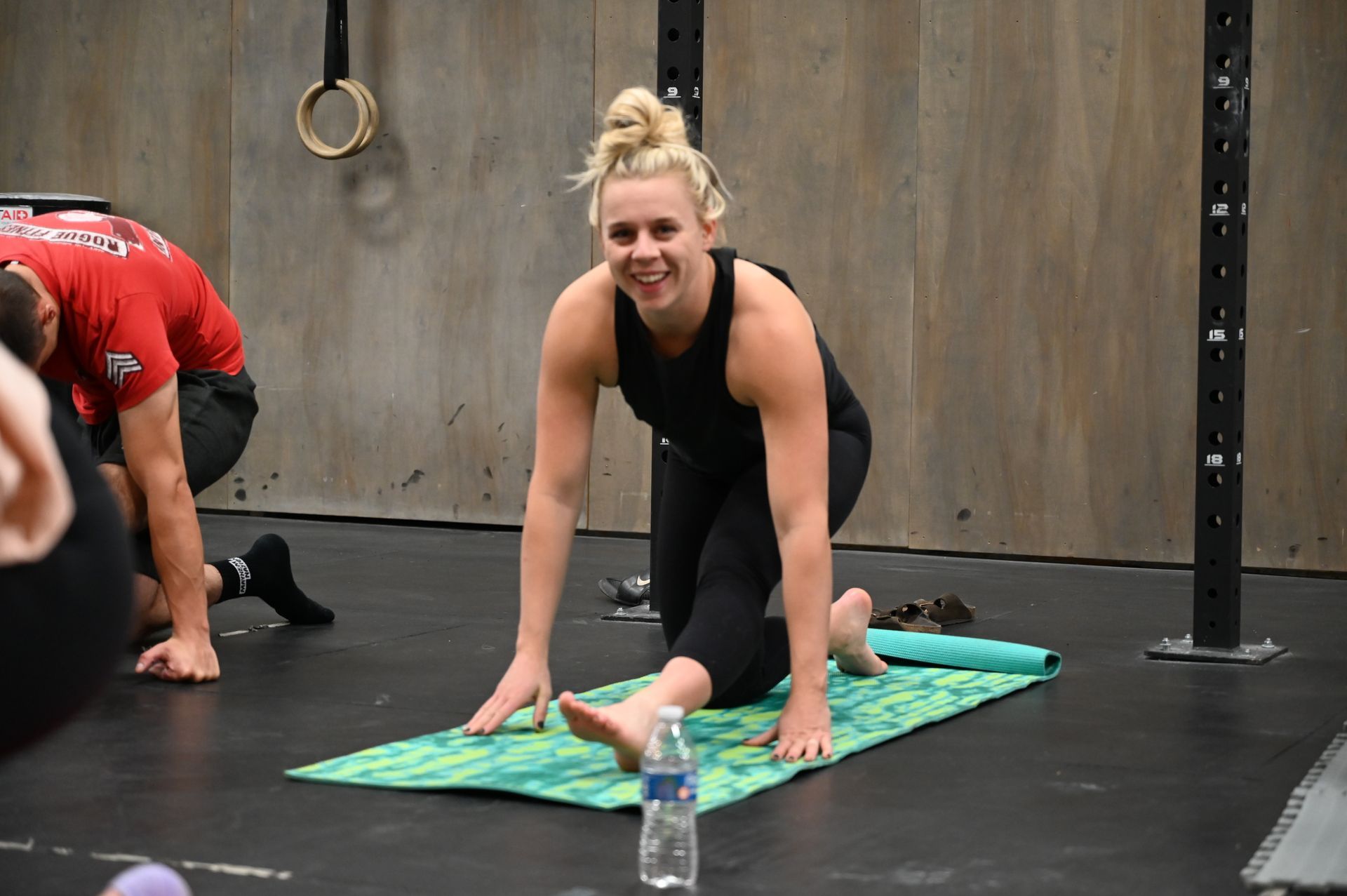 Woman smiles during a yoga stretch on a mat in a gym. She's in black activewear and has blonde hair.