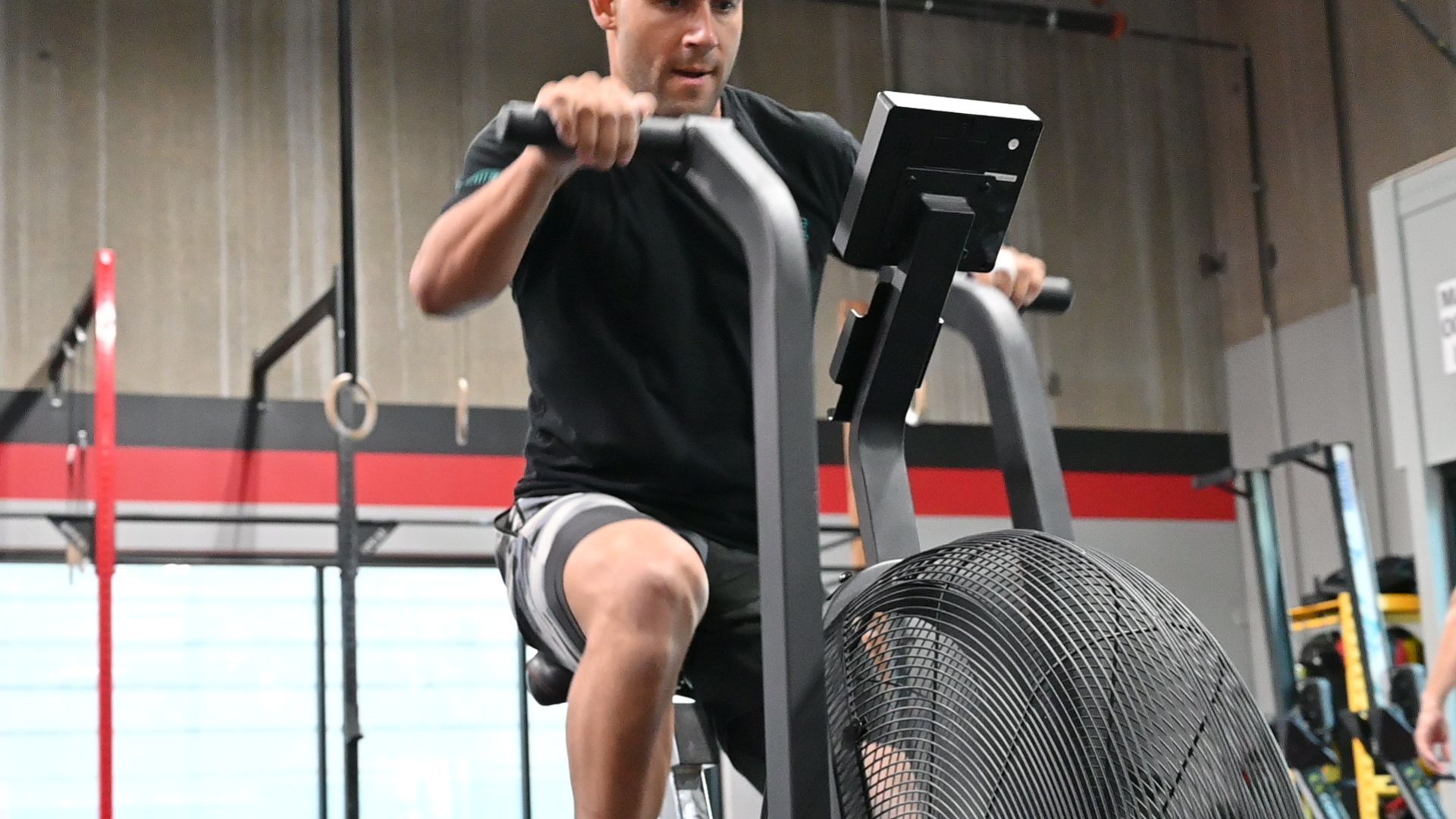 Man in gym, exercising on a stationary air bike. He is wearing a black shirt, shorts, and is focused.