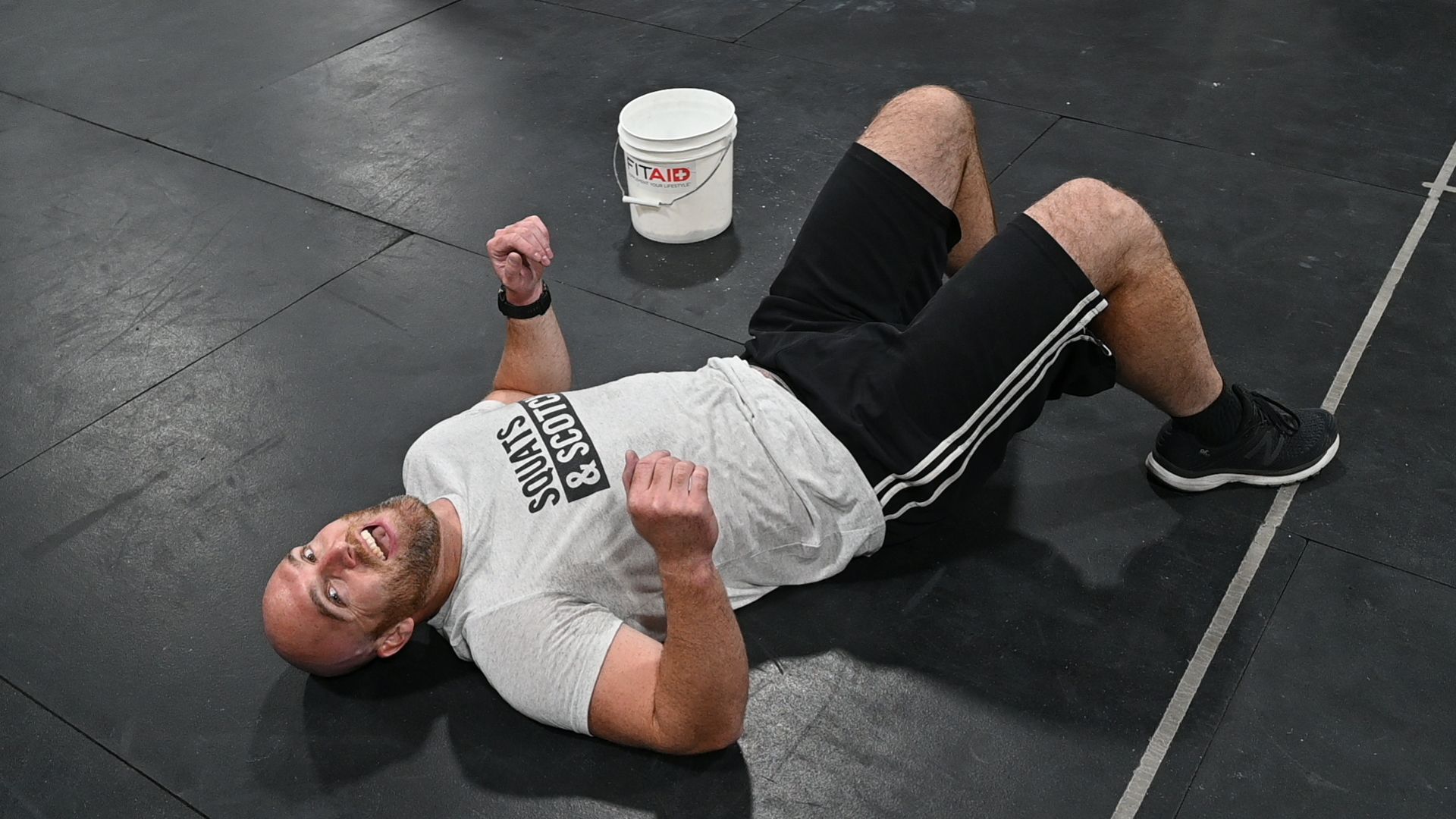 Man lying on back, arms up, legs bent, black shorts and shoes, white shirt, bucket nearby.