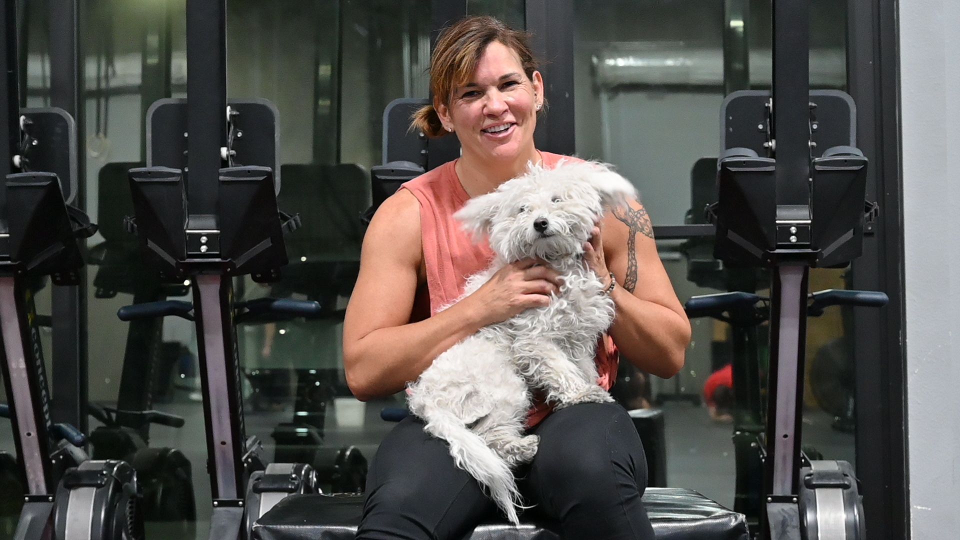 Woman in gym holding small, white fluffy dog; smiling. Row machines in background.