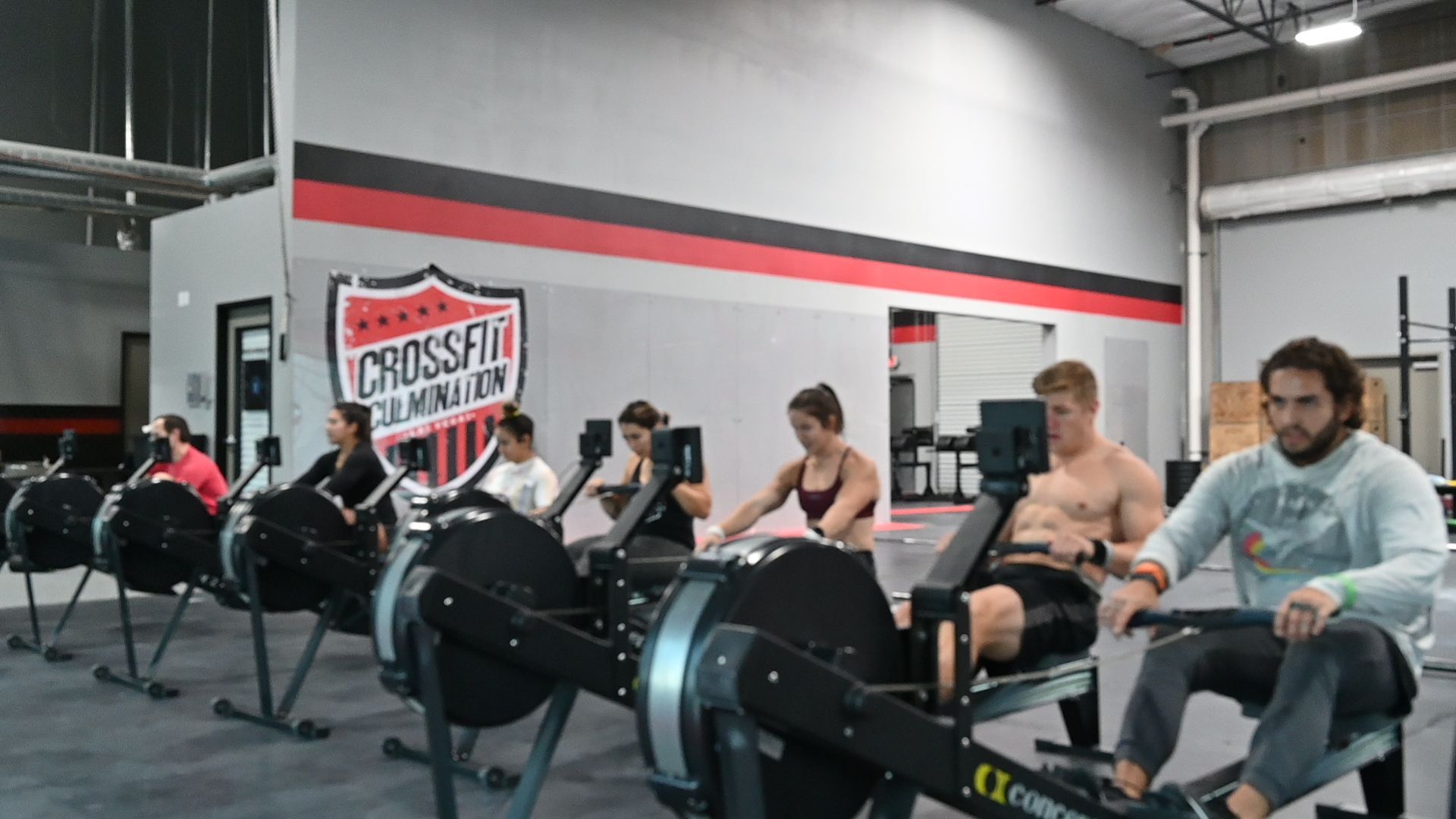 People rowing on Concept2 rowers in a CrossFit gym. Black machines, gray walls, red and black logo.