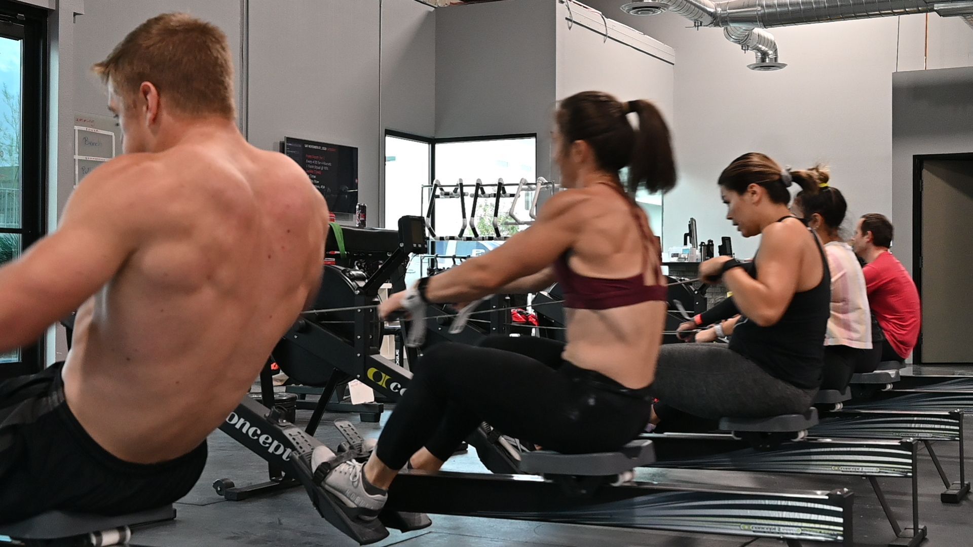 People rowing on exercise machines in a gym. Back view of a shirtless person rowing. Others are wearing workout clothes.