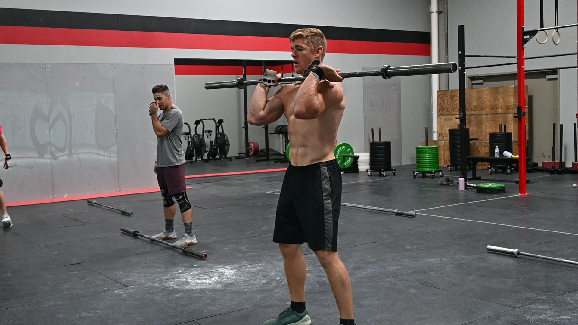 Man in gym holding barbell, preparing to do a front squat, other people in the background.