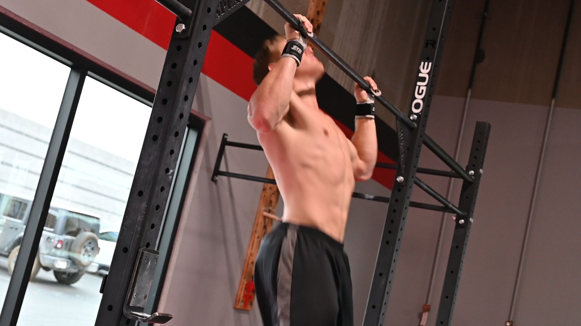 Man doing a pull-up on a black bar inside a gym, with wrist wraps, gray pants, and a bare torso.