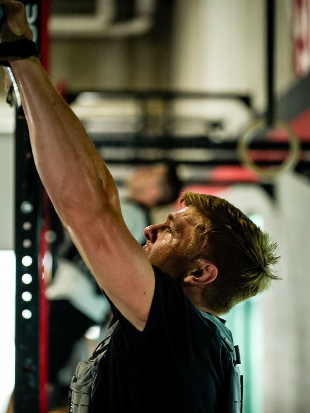Man, sweating, straining to hang from a pull-up bar, arm extended upwards. Gym setting.
