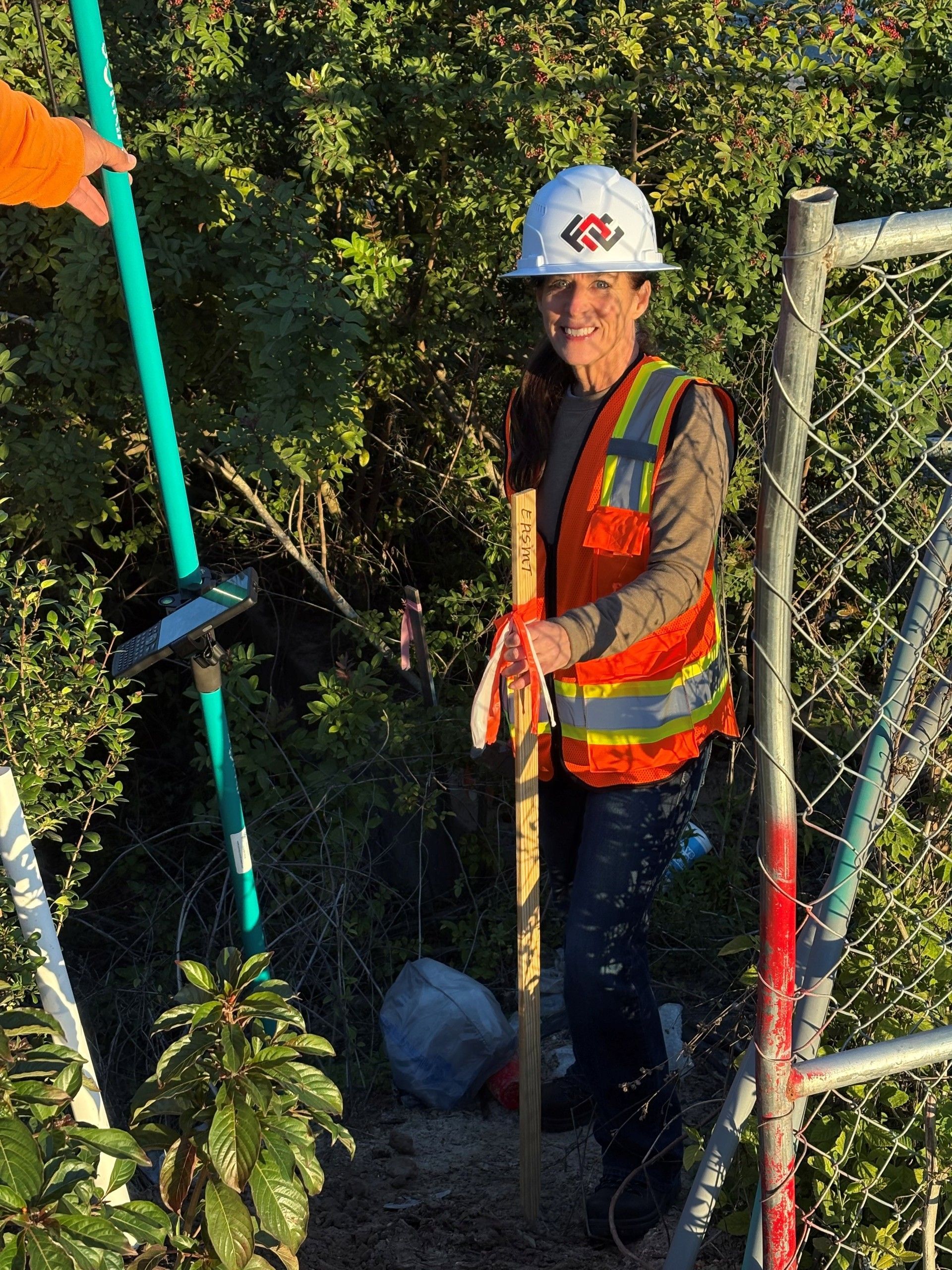 A person in a hard hat and high-visibility vest stands outdoors near a chain-link fence, holding a wooden survey stake.