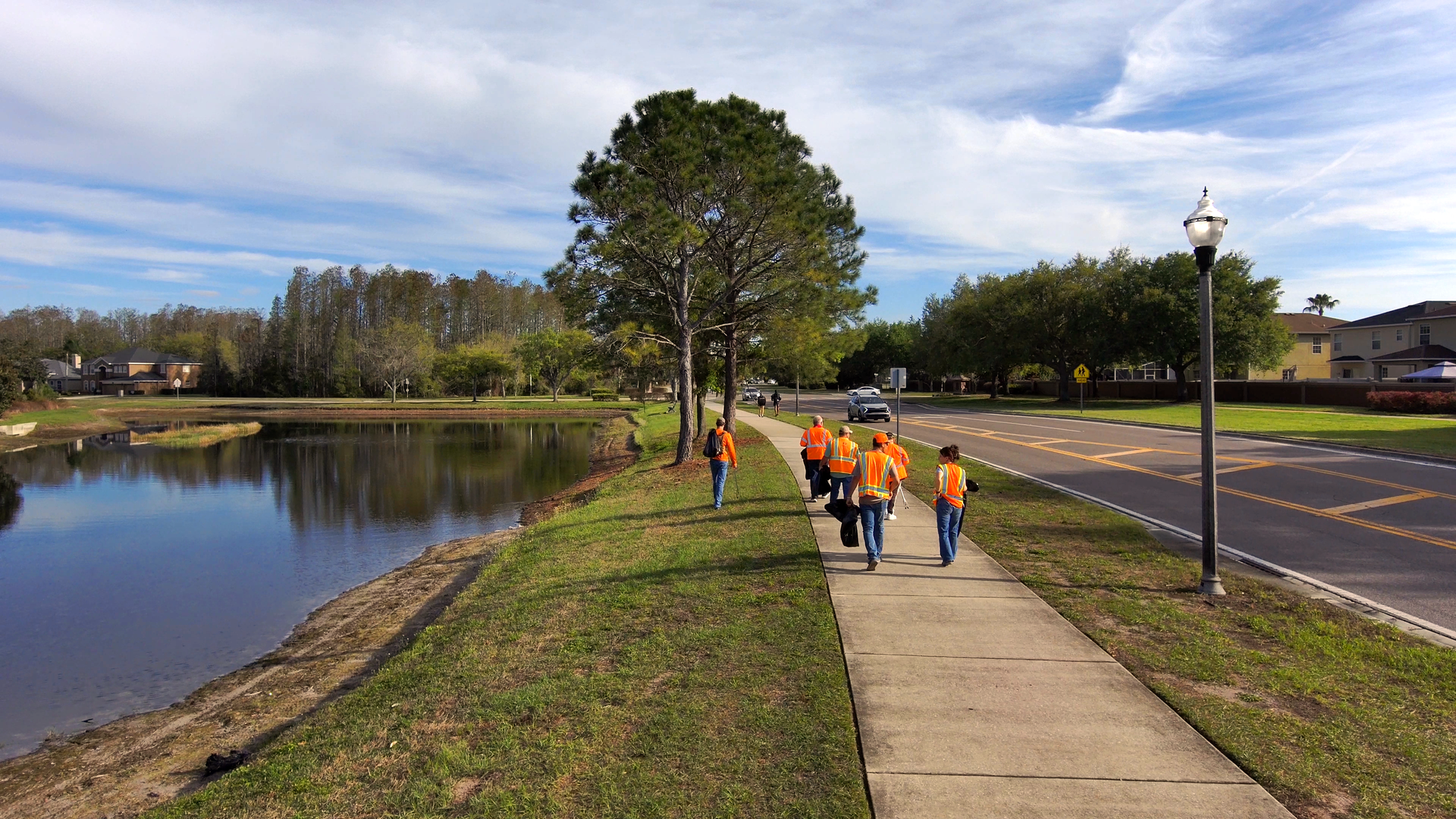 Children in orange shirts walking along a lakeside sidewalk beside a road and trees