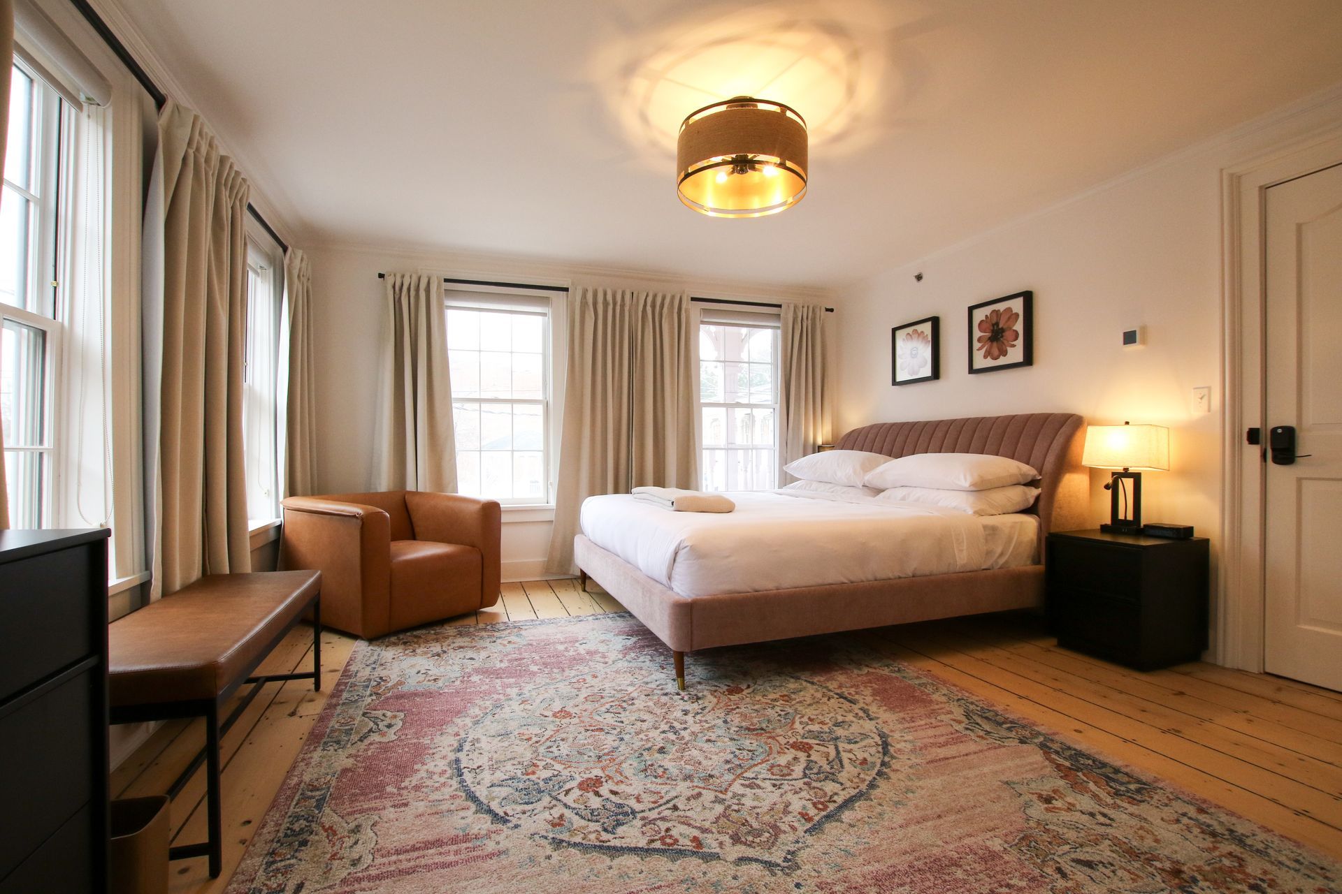 A large room with king size bed and four windows draped by white velvet curtains. Pink upholstery adorns the bed and carpet. A pendant light hangs over the bed.