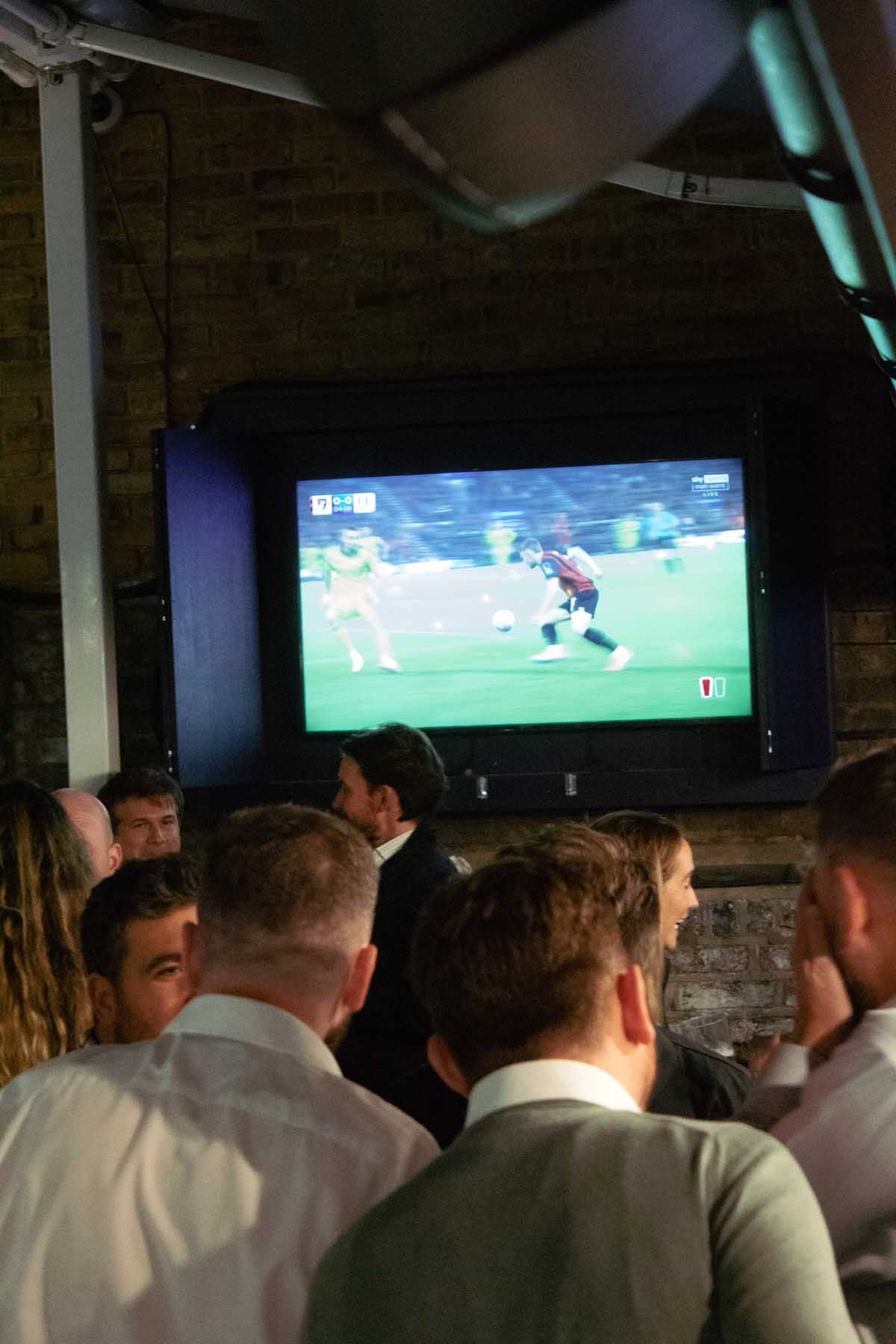 TV screen shows live football match in a pub with people watching a soccer game on a large screen.
