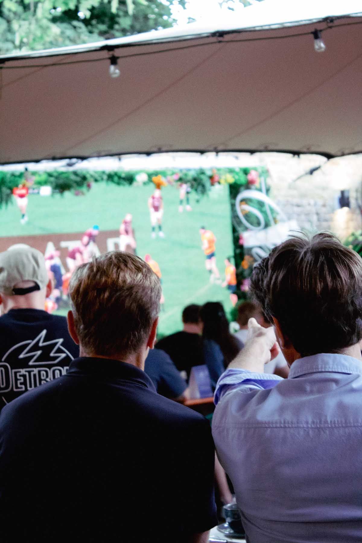 Two me sit at a table in a restaurant watching a rugby game on a large screen.