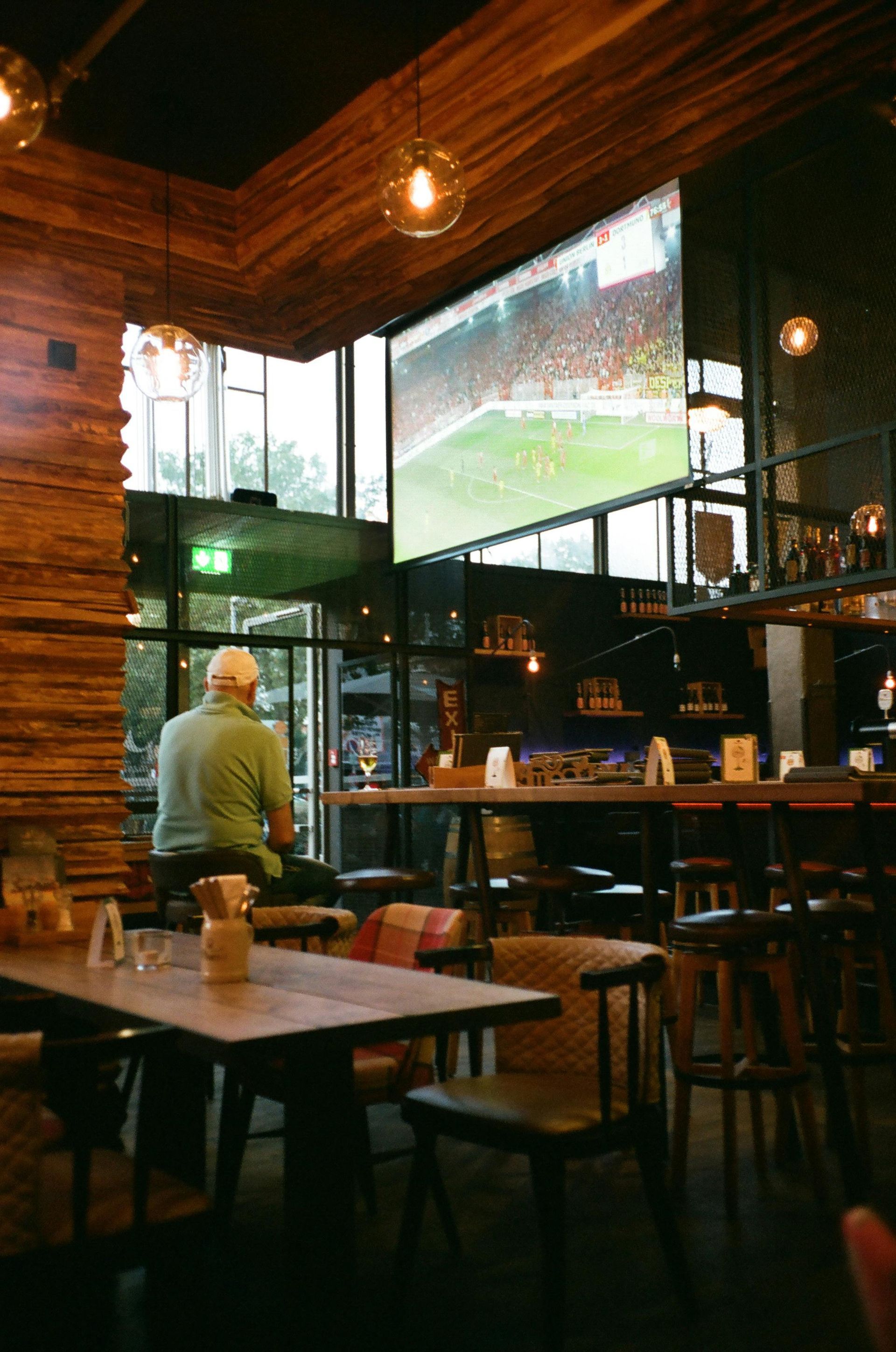 A man sits at a table in a restaurant watching a soccer game on a large screen.