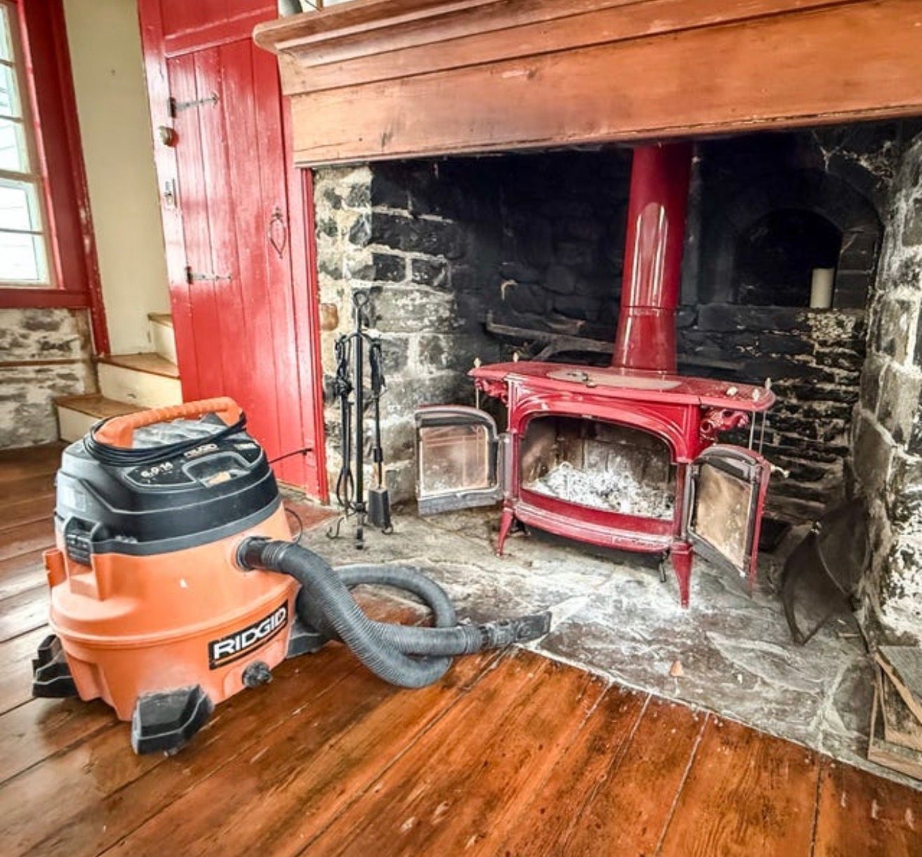 An orange shop vacuum sits on a hardwood floor in front of a stone fireplace with a red wood-burning stove inside.