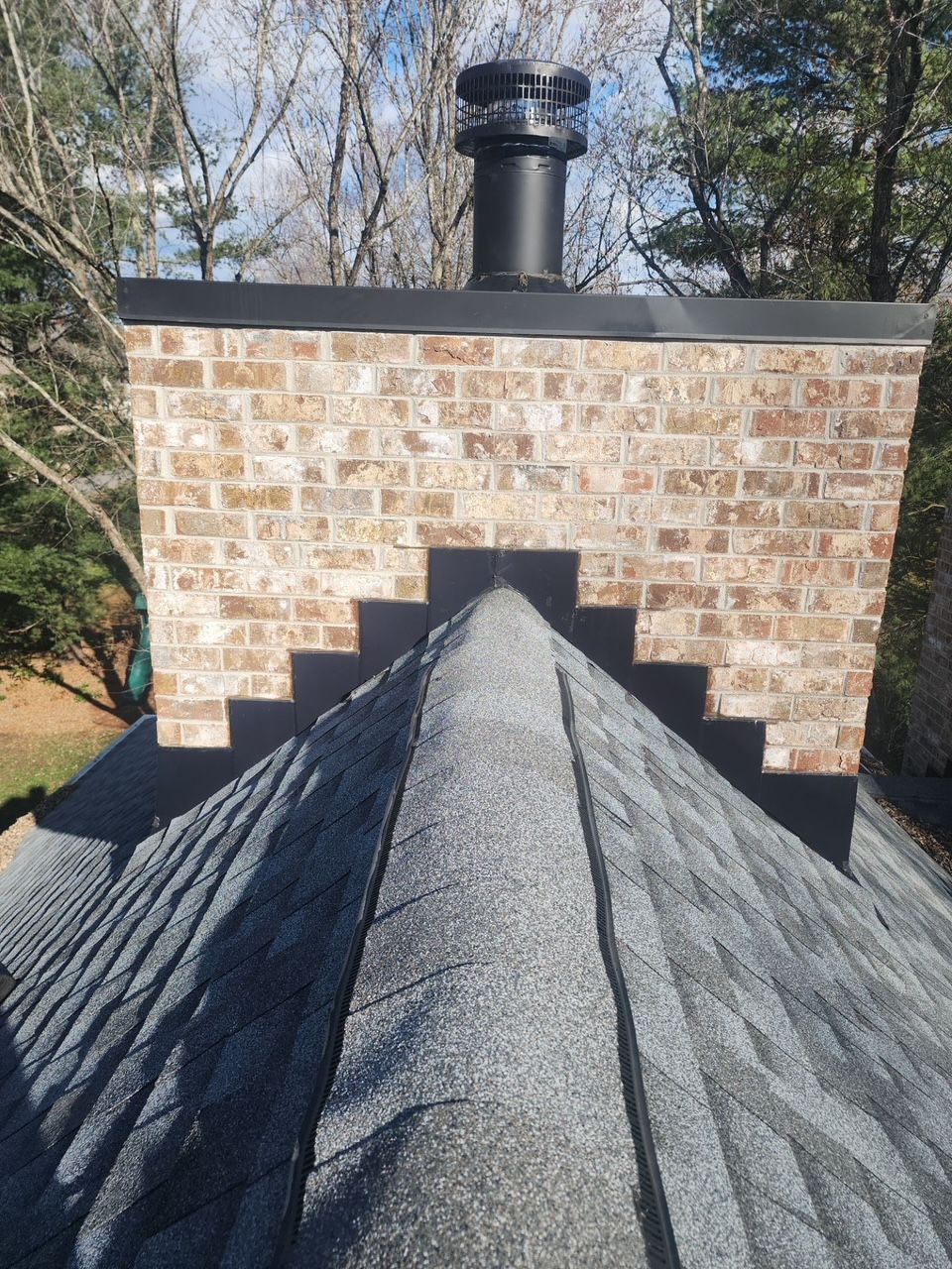 A brick chimney with a metal cap centered behind the ridge of a shingled roof.