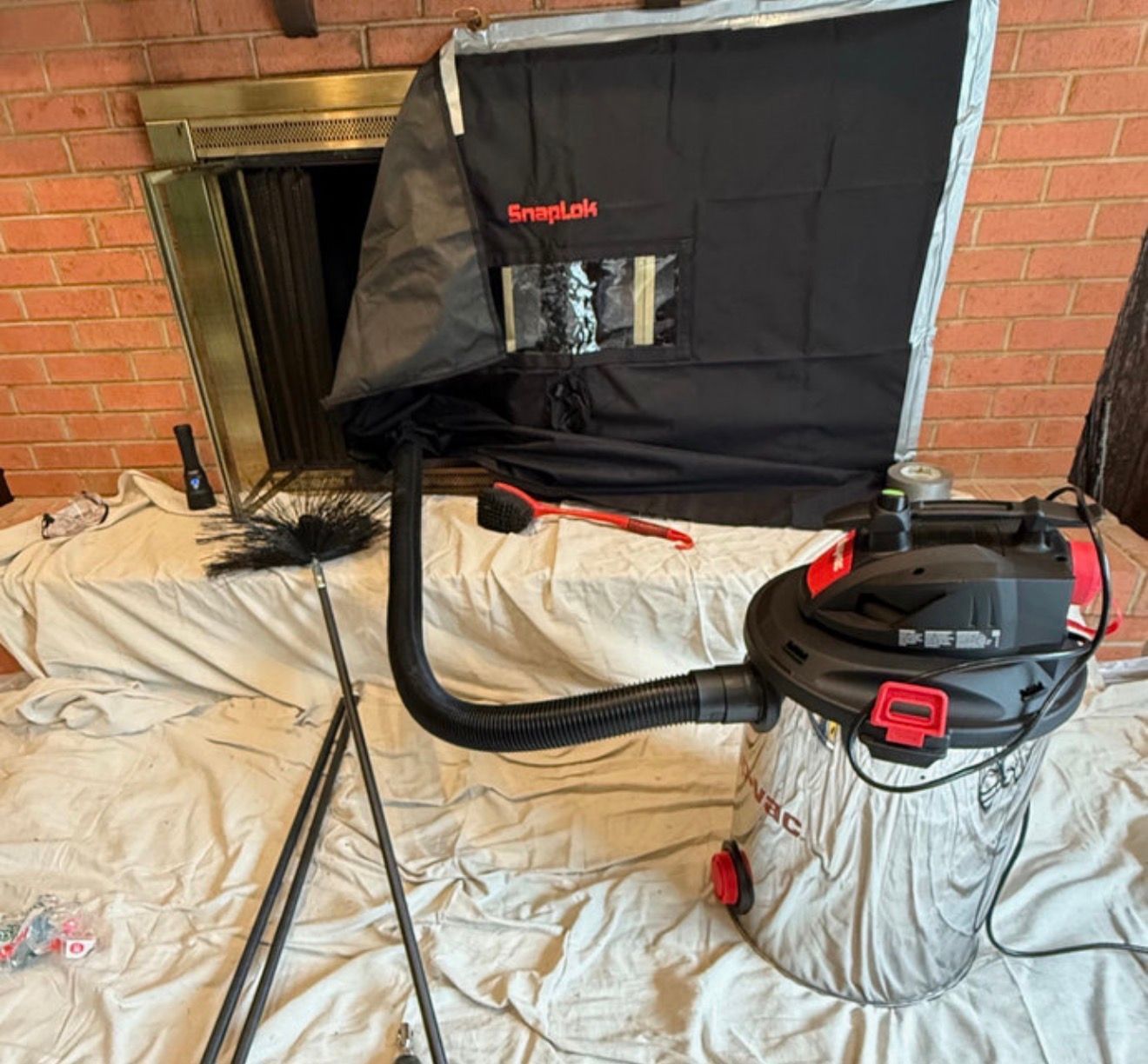 A fireplace cleaning setup with a vacuum, brush, and a soot-containment cover on a drop cloth in front of a brick fireplace.