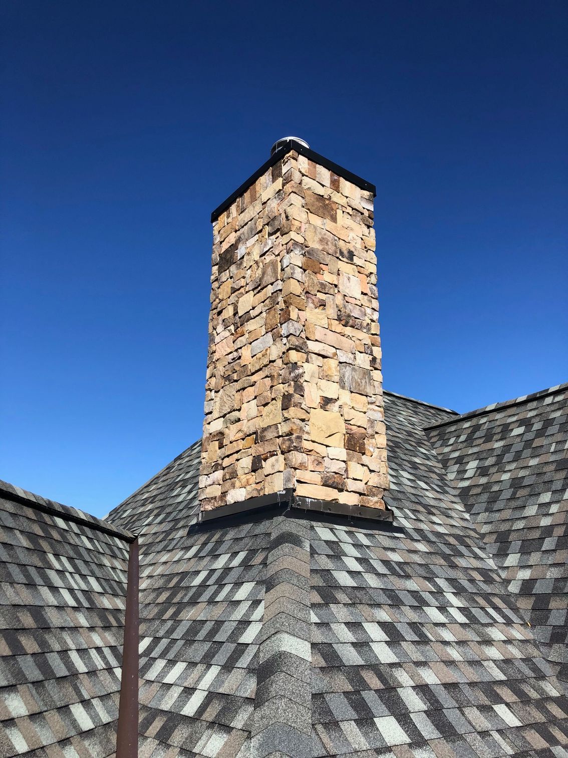 A tall, stone-clad chimney rises from a pitched roof with dark, multi-toned asphalt shingles against a clear blue sky.