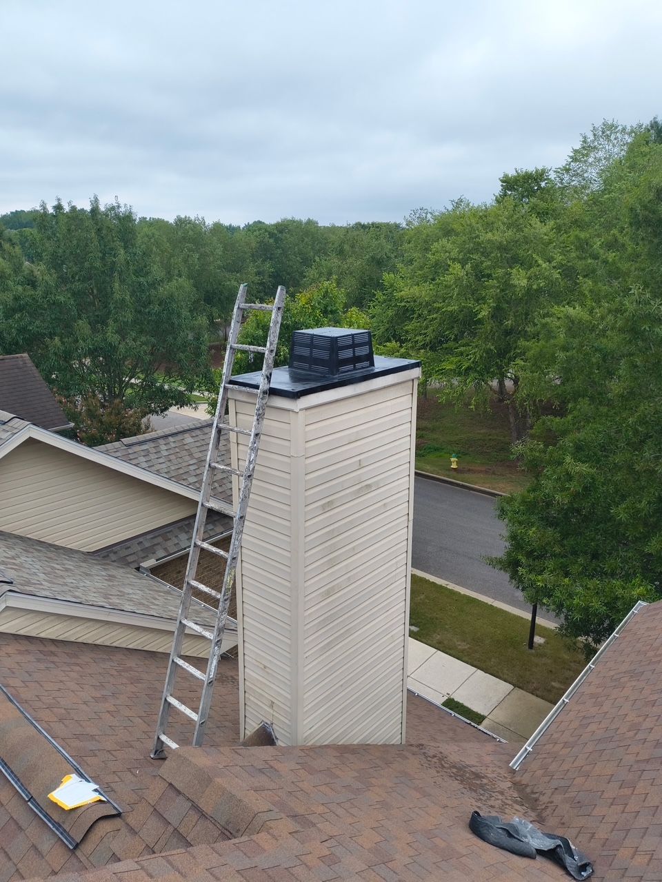 A tall, beige, sided chimney stands on a shingled roof with a metal extension ladder leaning against its side.