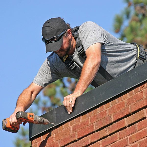 A worker wearing a safety harness and cap uses a power drill to install metal flashing onto a brick wall.