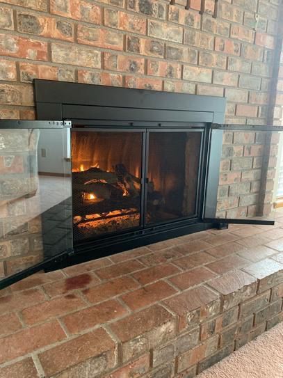 A brick fireplace with an open black glass-paneled door, revealing a glowing fire inside.