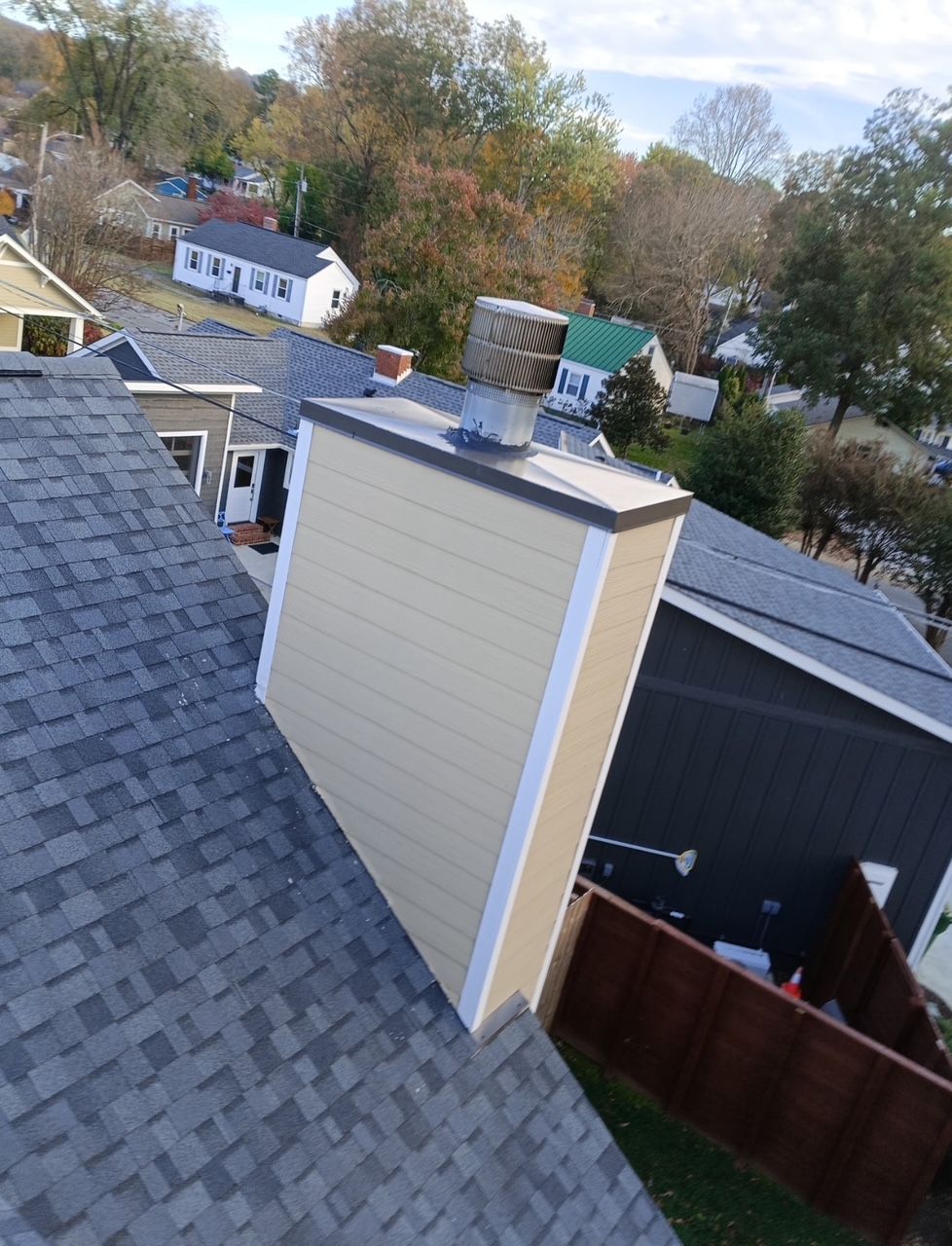 A view from a shingled roof looking down at a beige chimney with a metal cap, surrounding houses, and trees.