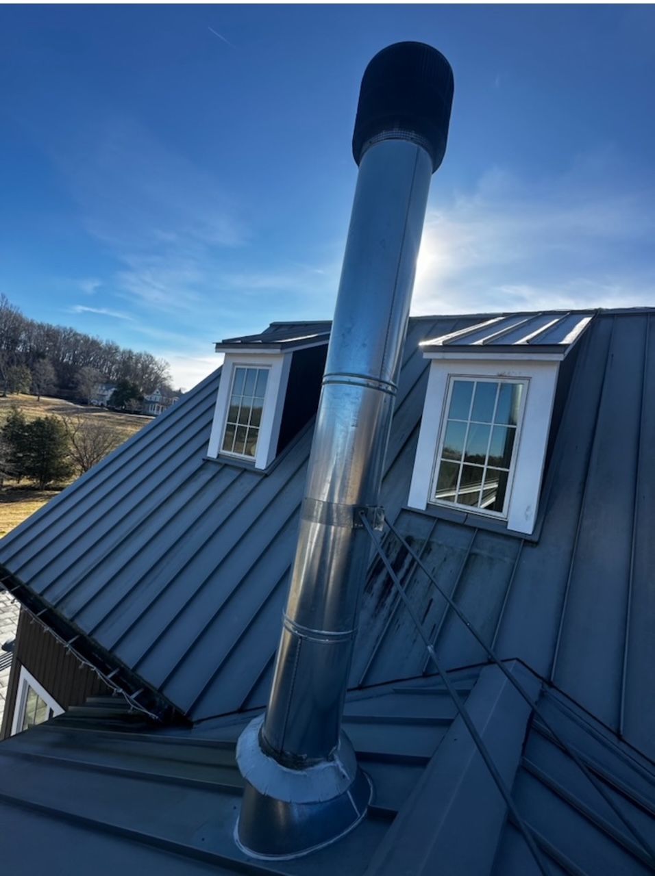 A tall, stainless steel chimney pipe extending from a dark metal roof, situated between two small dormer windows.