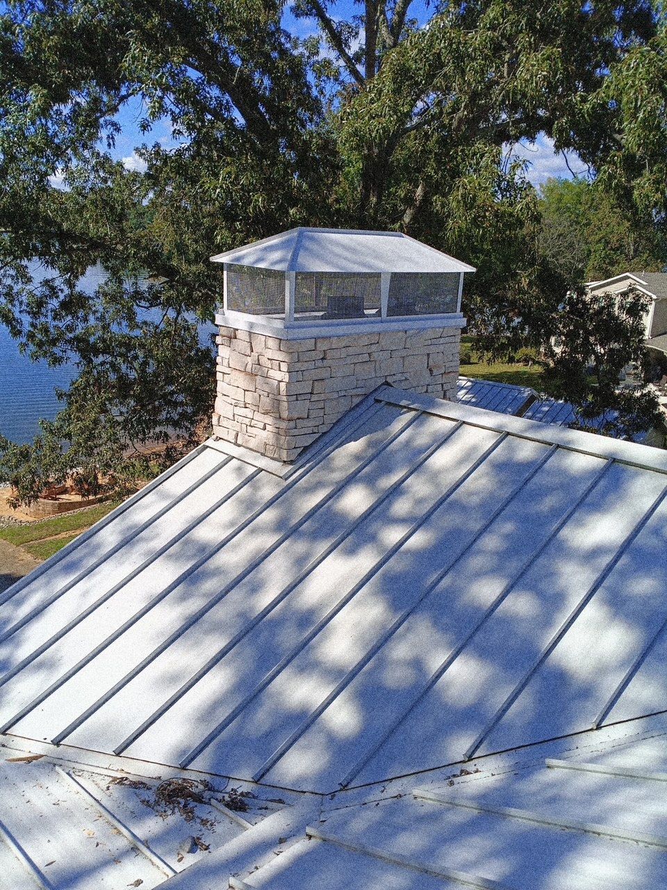 A light-colored stone chimney with a metal cap sits on a silver standing-seam metal roof near trees and water.