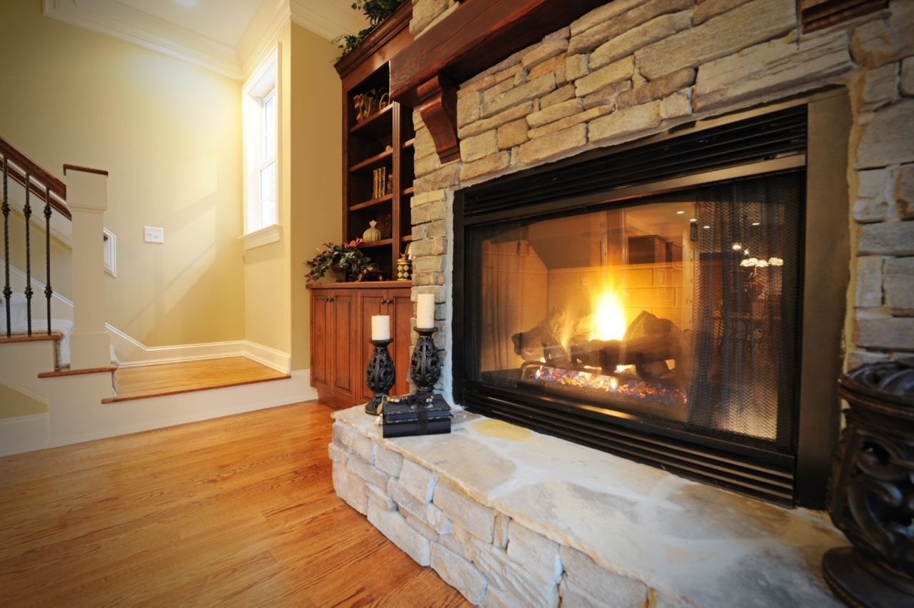 A stone fireplace with a glowing fire sits next to a wooden bookcase and staircase in a home interior.
