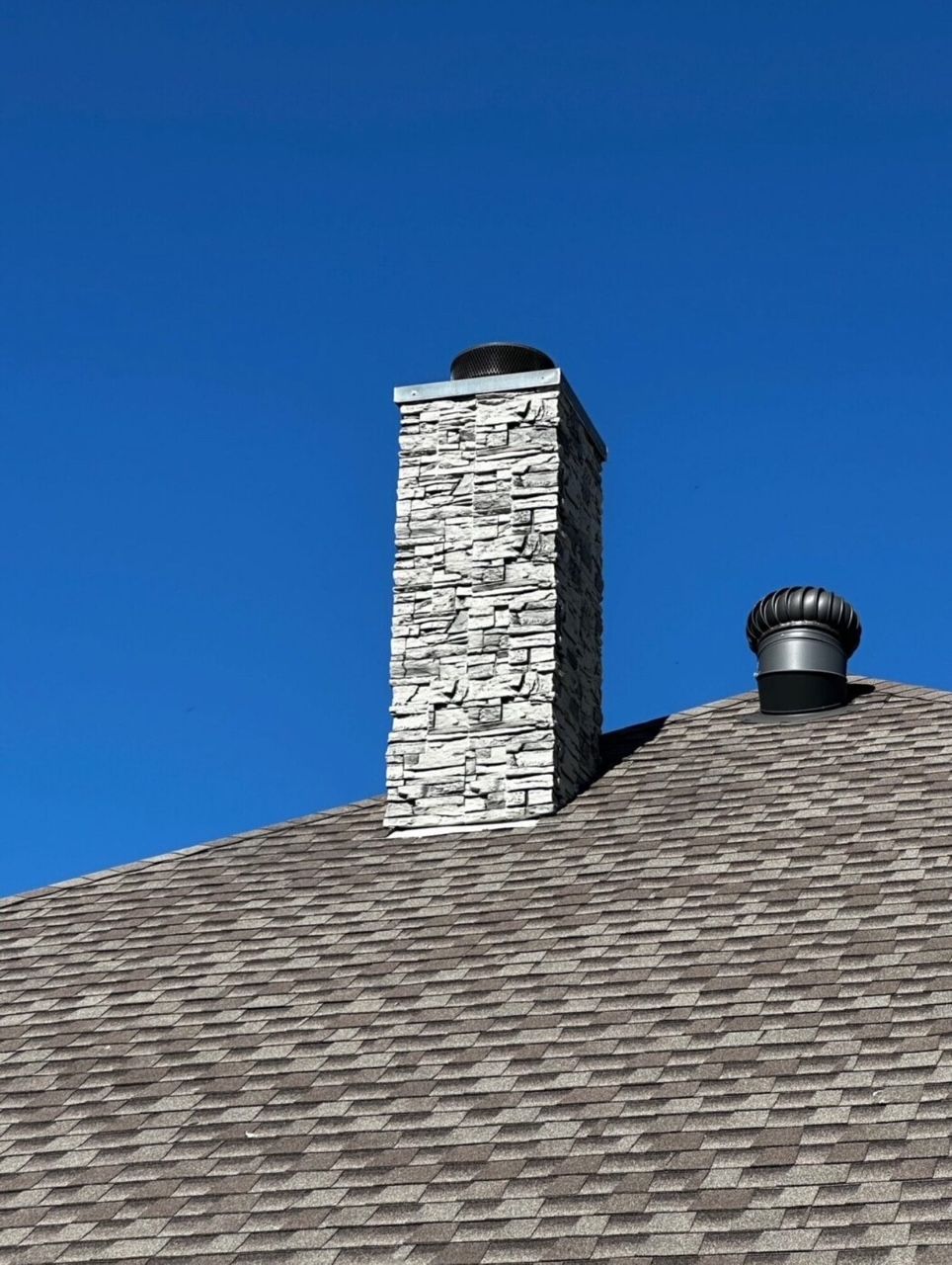 A stone chimney and a metal roof vent protruding from a brown shingled roof against a bright blue sky.