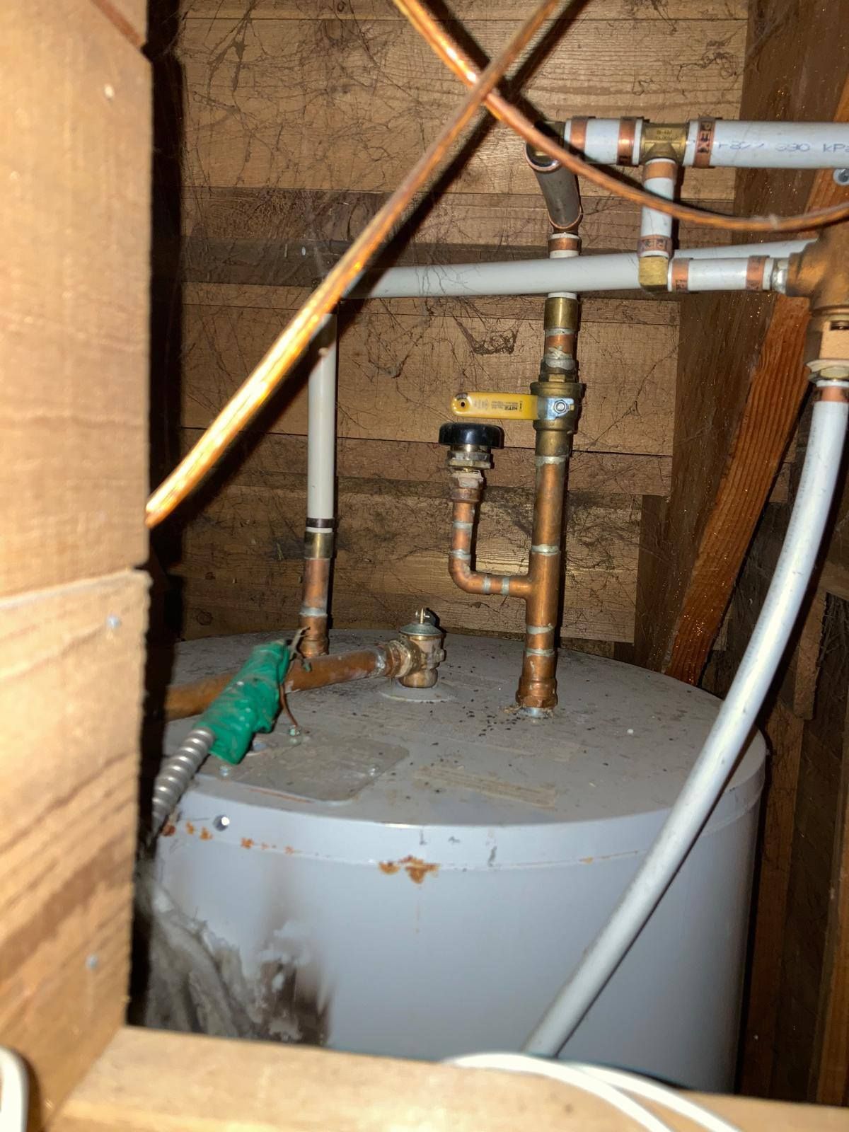 A gray water heater in an attic, connected to white PEX piping, copper fittings, and a yellow-handled shut-off valve.