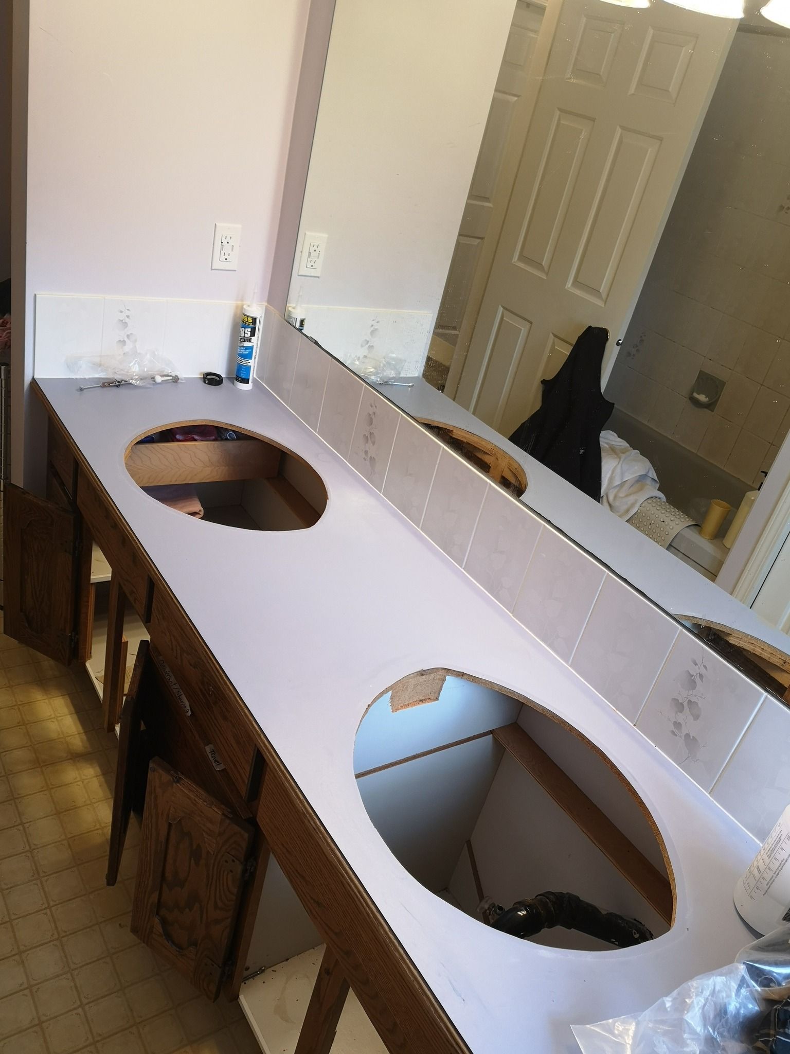 A long, white bathroom vanity top with two oval sink cutouts sits on a wooden cabinet, reflected in a large wall mirror.