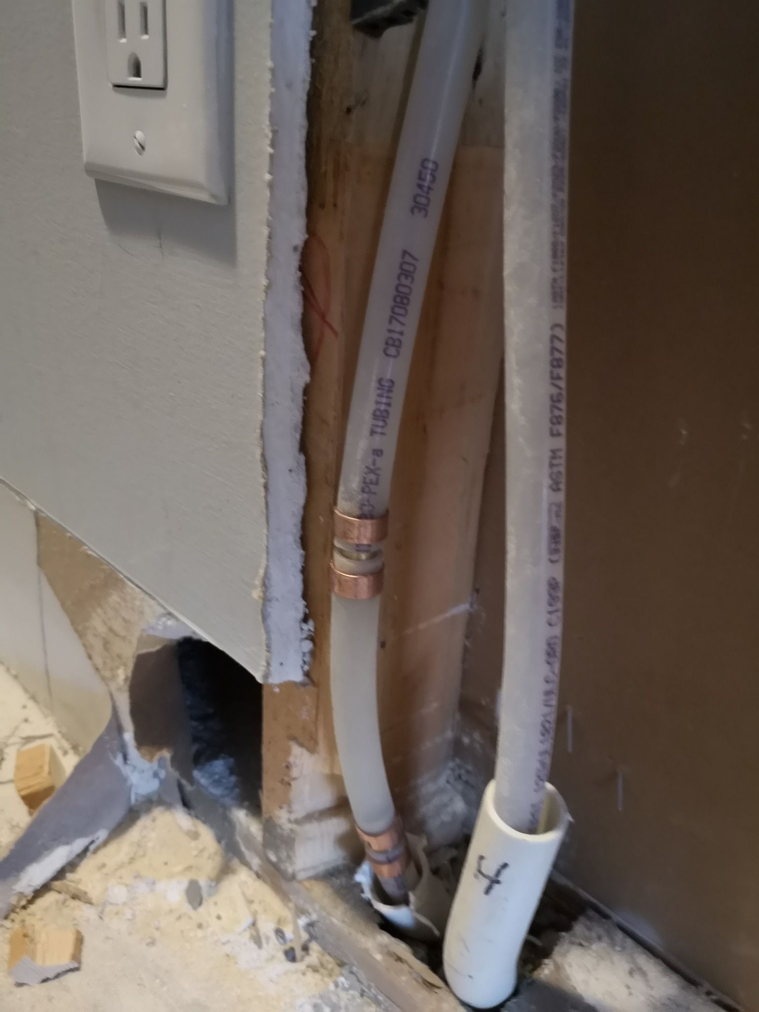 Two white PEX water pipes with copper couplings inside an open wall cavity near a power outlet.