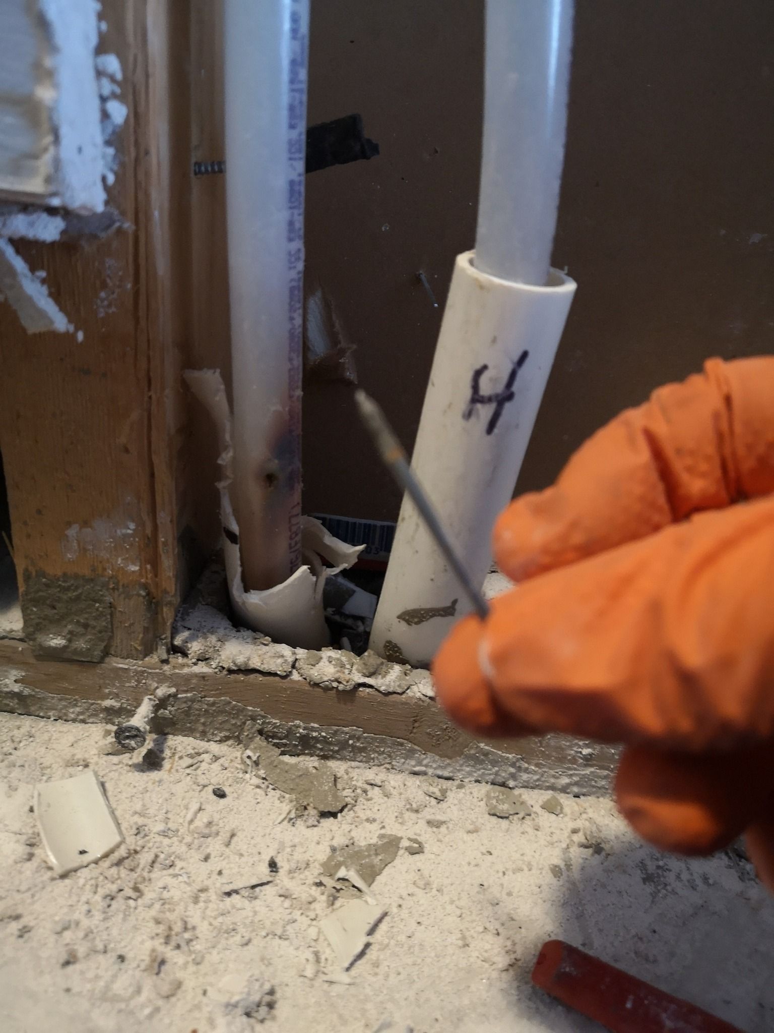 A gloved hand holds a nail that has punctured a white pipe inside a wall.
