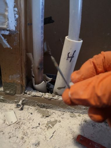 A gloved hand holds a nail that has punctured a white pipe inside a wall.