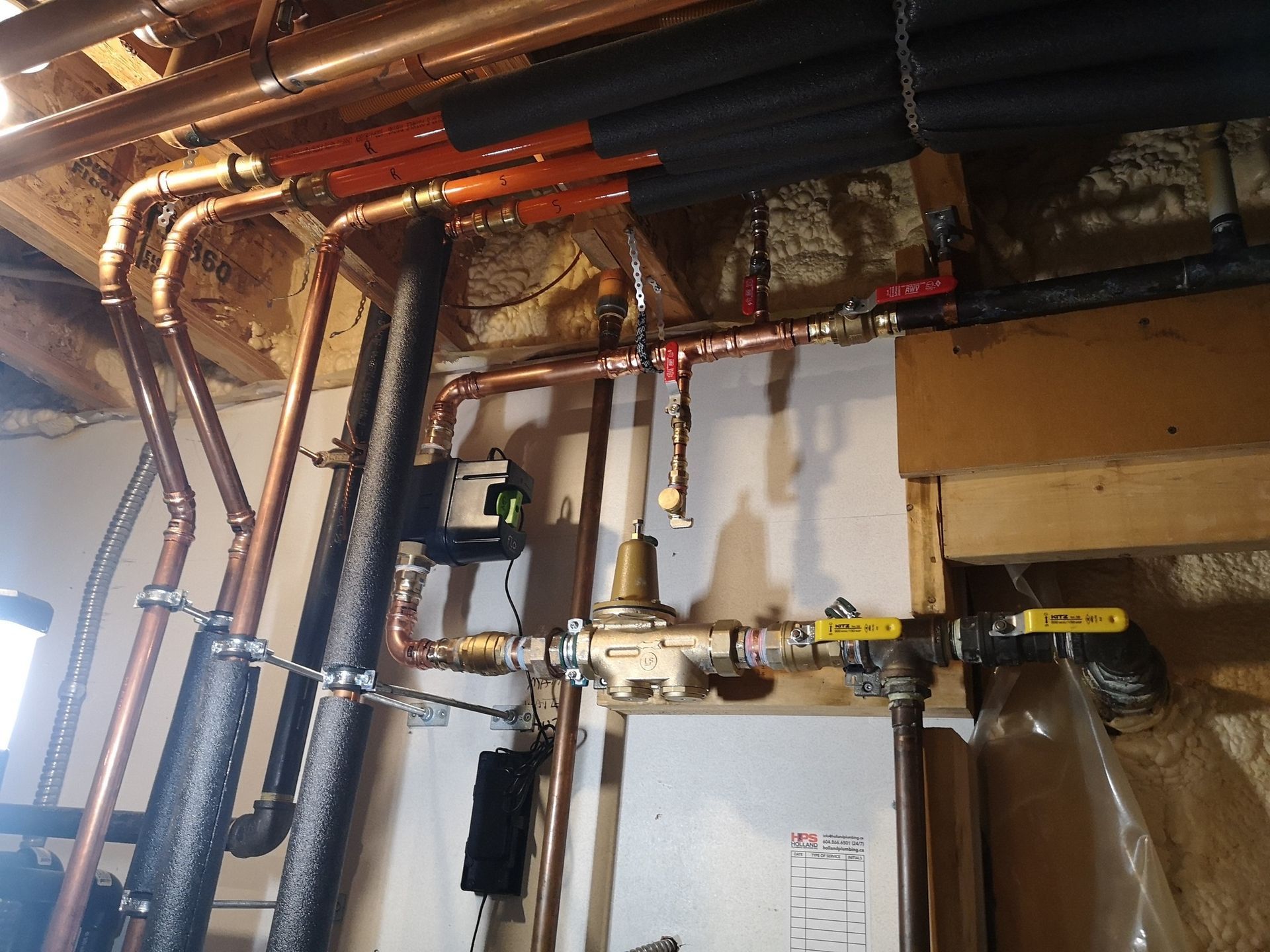 Residential copper piping system attached to wooden joists, featuring a pressure regulator, valves, and black insulation.