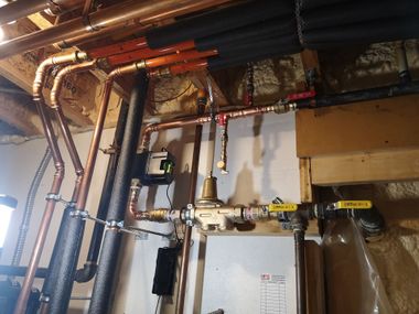 Residential copper piping system attached to wooden joists, featuring a pressure regulator, valves, and black insulation.