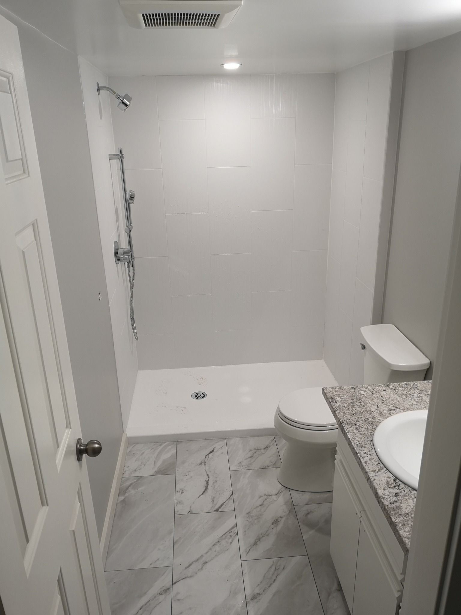 A small, modern bathroom with a white walk-in shower, a toilet, and a vanity with a granite countertop and marble floor.