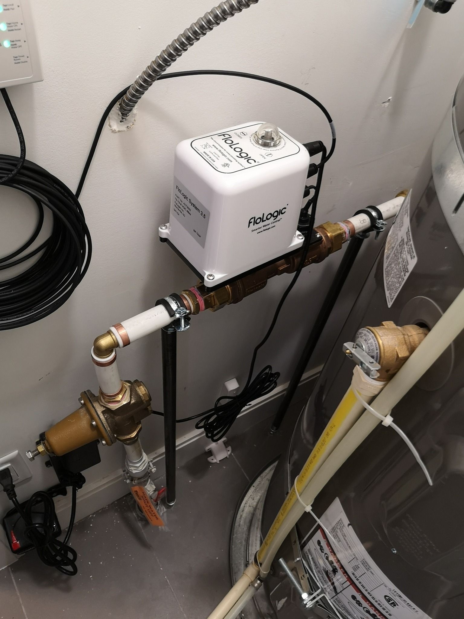 A white electronic water shut-off valve installed on plumbing pipes next to a water heater in a utility room.