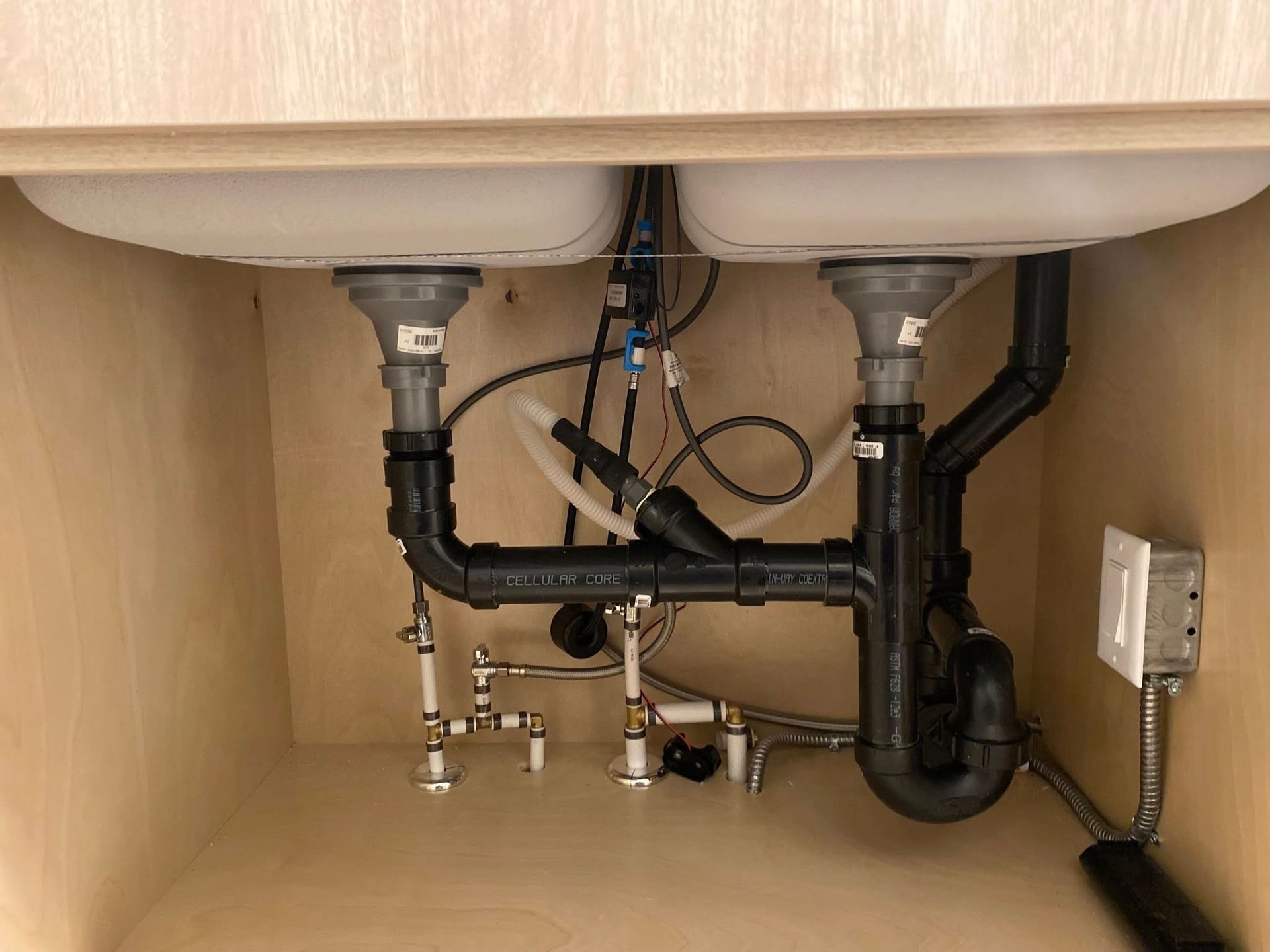 Under-sink cabinet view showing black PVC drain plumbing for a double-basin sink, water supply lines, and an electrical box.