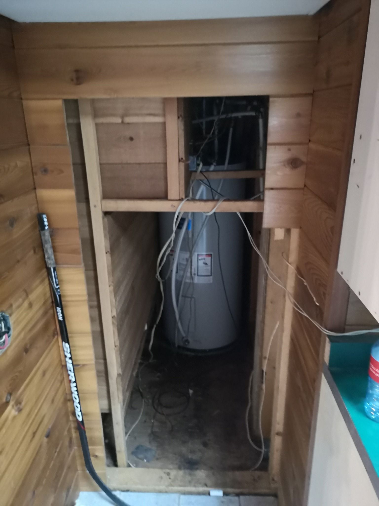 A utility closet with wooden walls containing a large water heater and exposed wiring in a narrow opening.